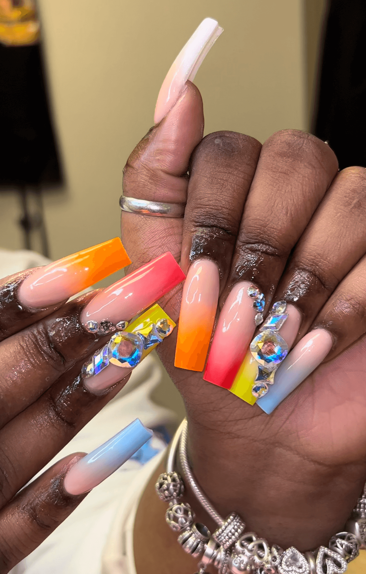 Close-up of two hands with colorful, decorated acrylic nails, some with rhinestones and gradient designs, against a blurred background.