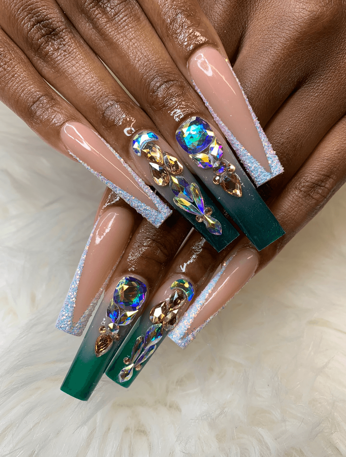 Close-up of long, decorated nails with rhinestones, gold accents, and a mix of pink and green colors, on dark skin.