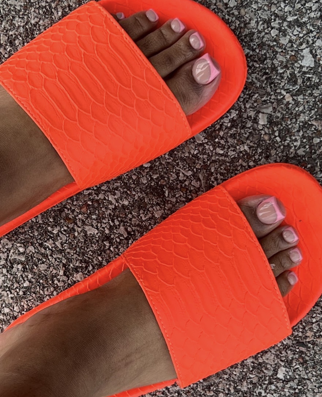 Person wearing bright orange slide sandals with textured straps, standing on a rough gray surface.