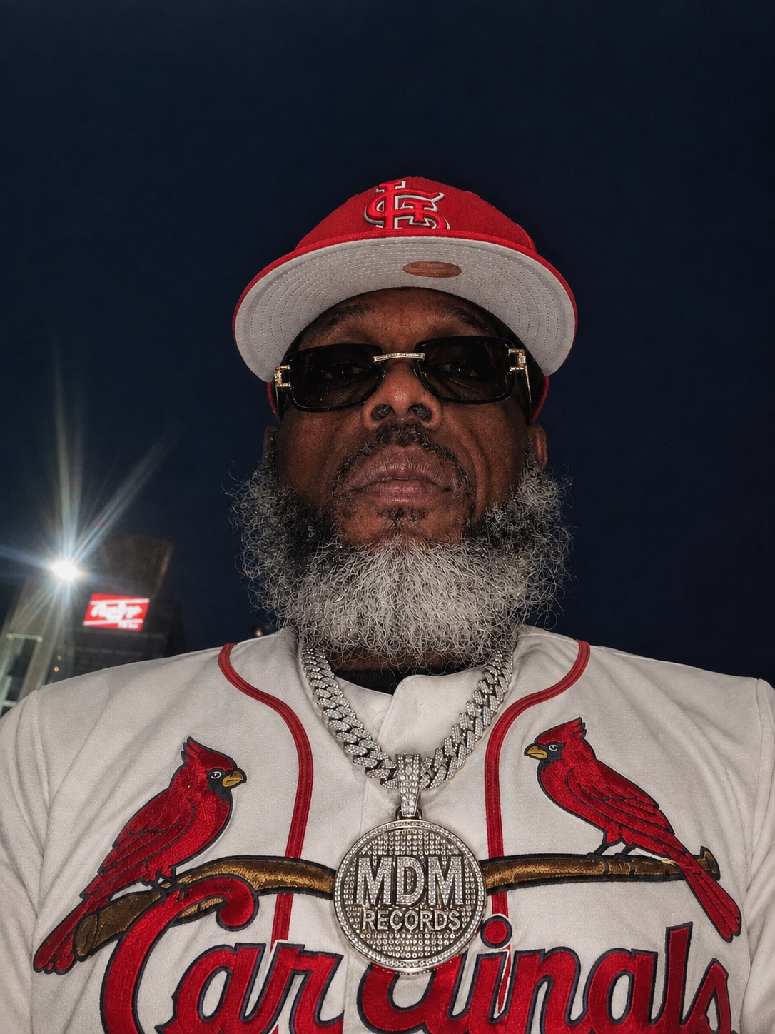 Man with sunglasses, gray beard, wearing a red and white cap, white jacket with red cardinal logos, and a large chain with a pendant.