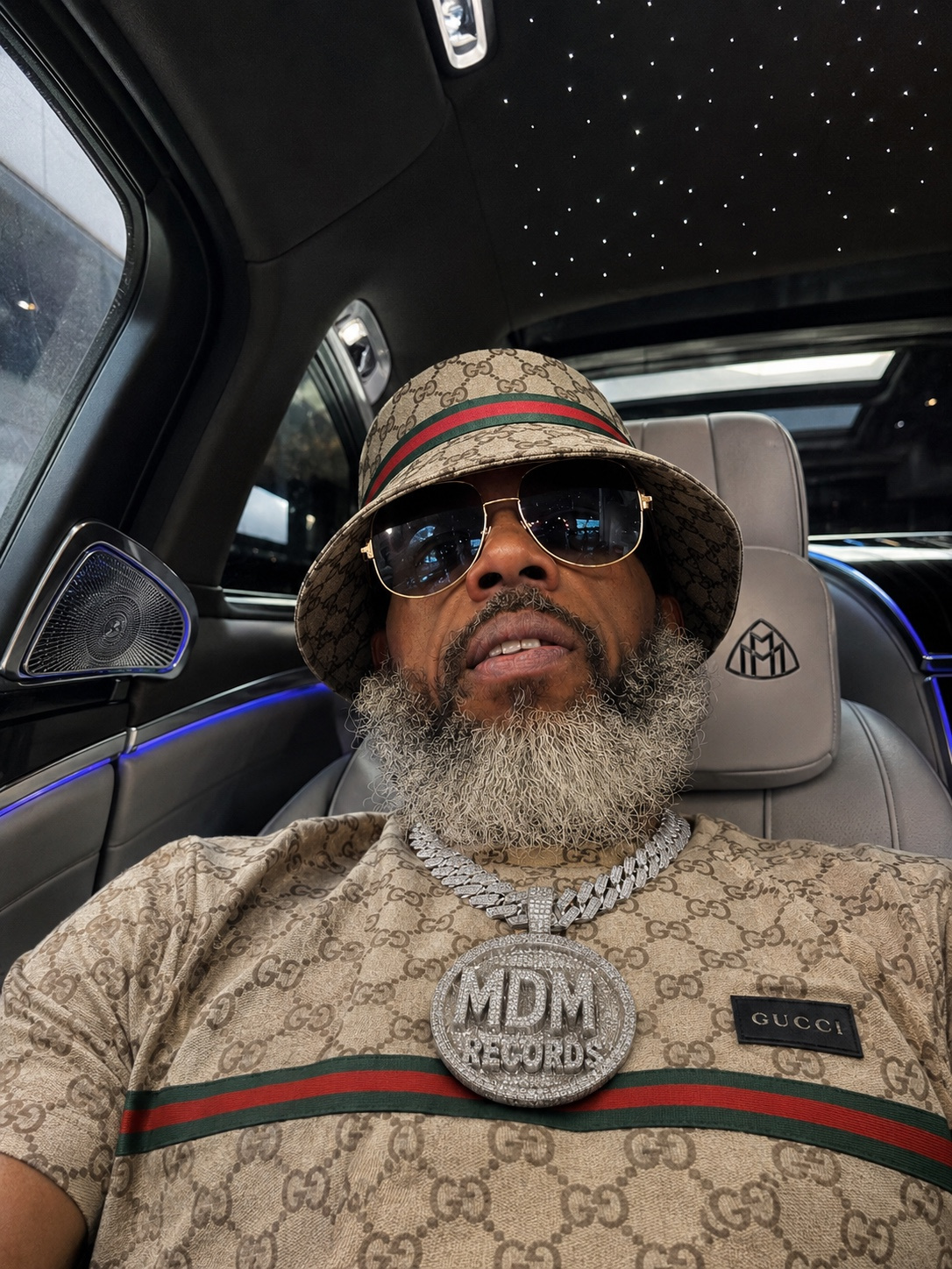 Man with sunglasses and a hat sitting in a car with a starry ceiling, wearing a Gucci shirt and large chain.