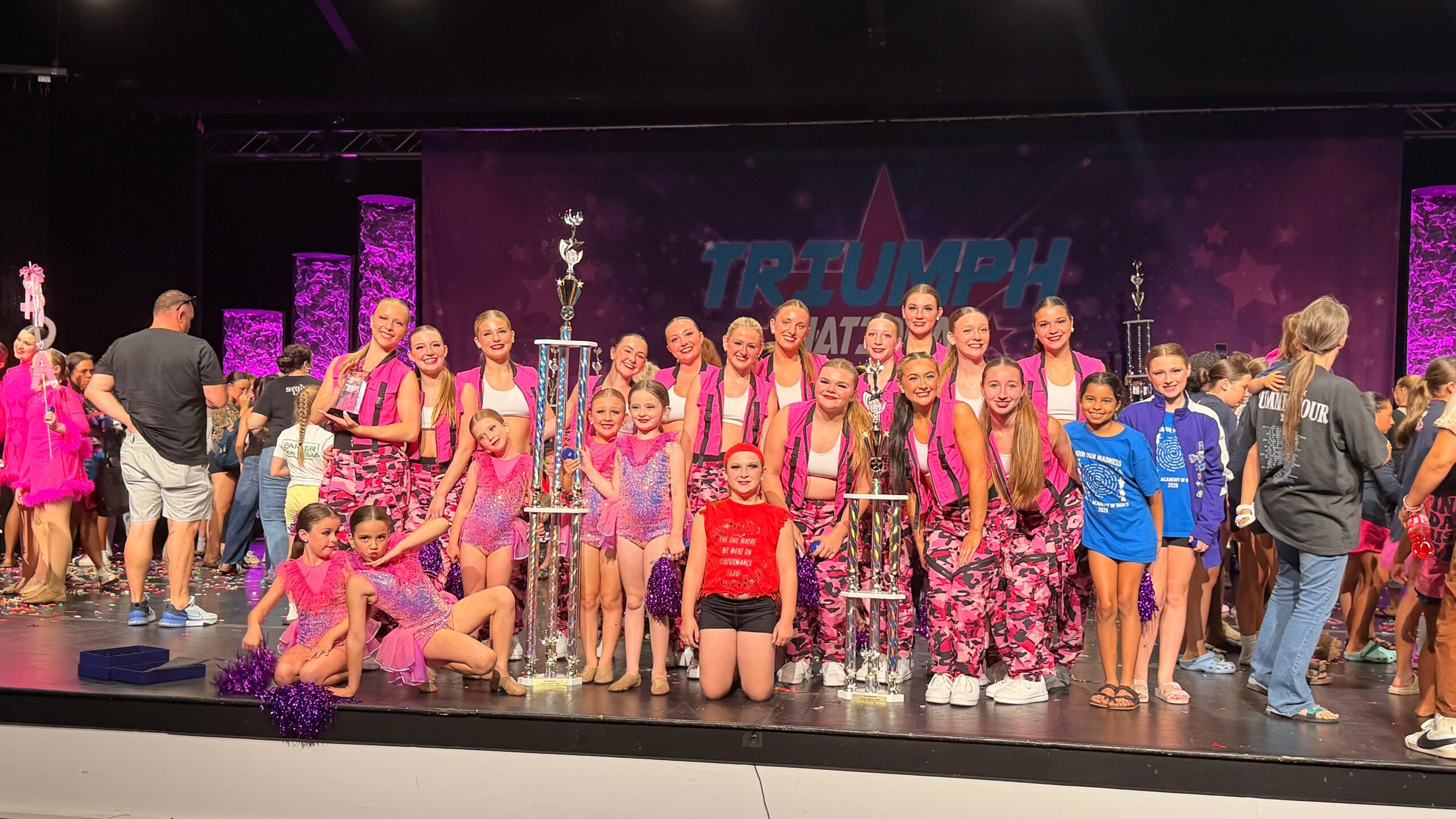 Group of children and adults on stage with trophies, some in colorful costumes, in a dance competition setting.