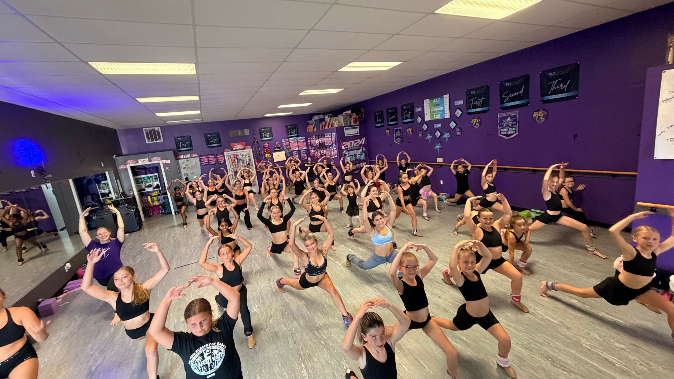 Group of people practicing dance or exercise in a studio with purple walls and mirrors, some stretching or posing.