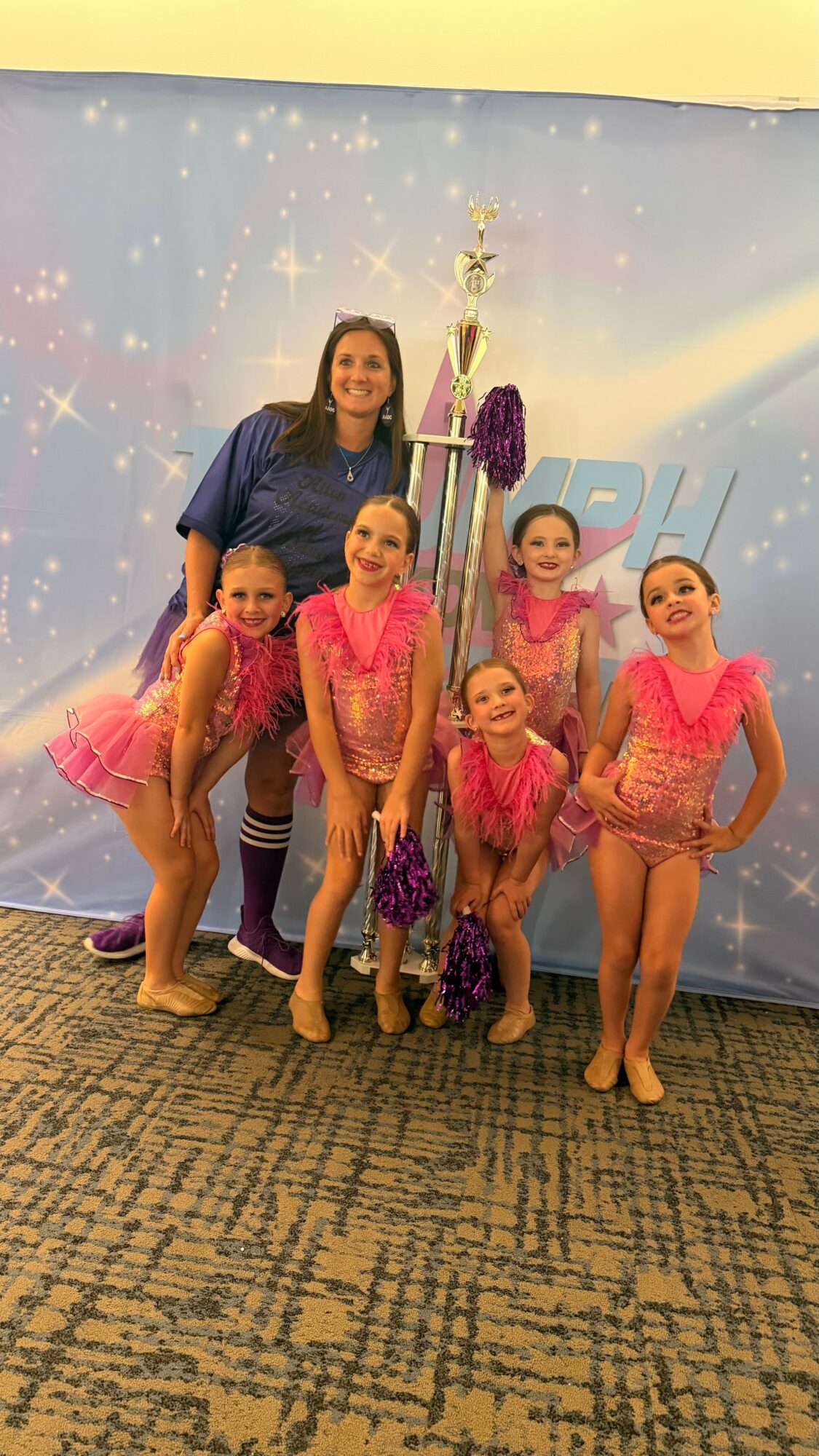 Group of five young girls in pink costumes with a woman, standing in front of a blue backdrop with sparkles and a trophy, on a carpeted floor.