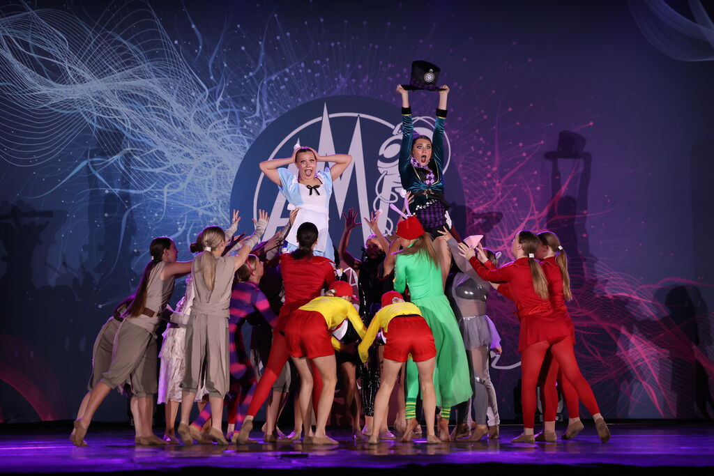 Group of performers on stage with two central figures, colorful costumes, and a backdrop with abstract designs.
