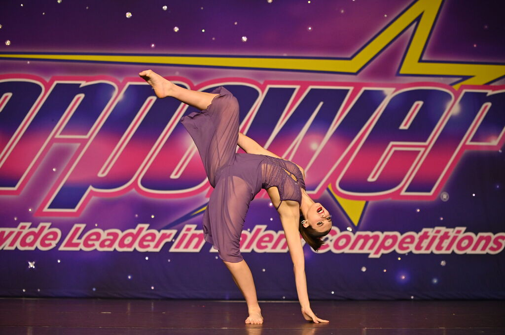 A person performs a handstand on stage with a colorful backdrop and text, wearing a purple outfit.