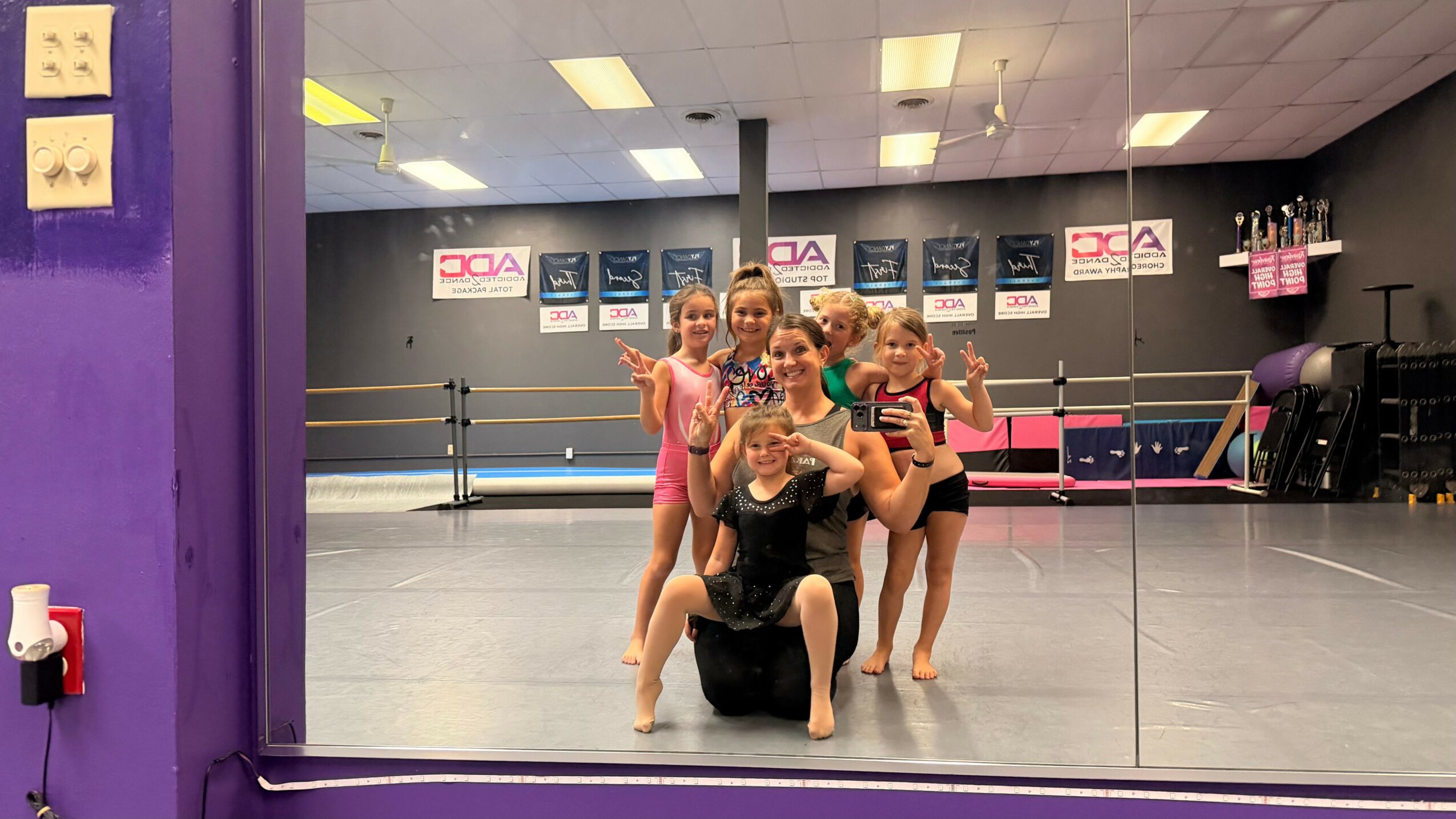 Group of young girls and an adult in a dance studio taking a mirror selfie, with dance posters on the wall.