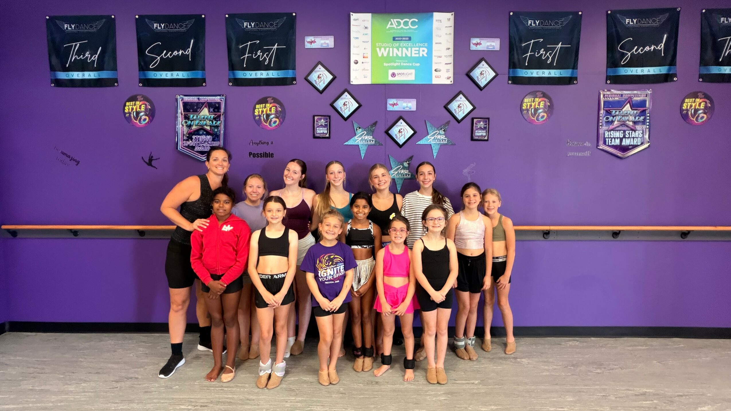 Group of young dancers in dance studio with purple wall, awards, and certificates displayed, posing for a photo.