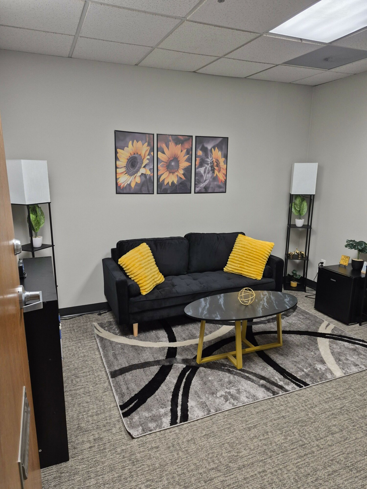 Living room with black sofa, yellow pillows, glass coffee table, and wall art of sunflowers.