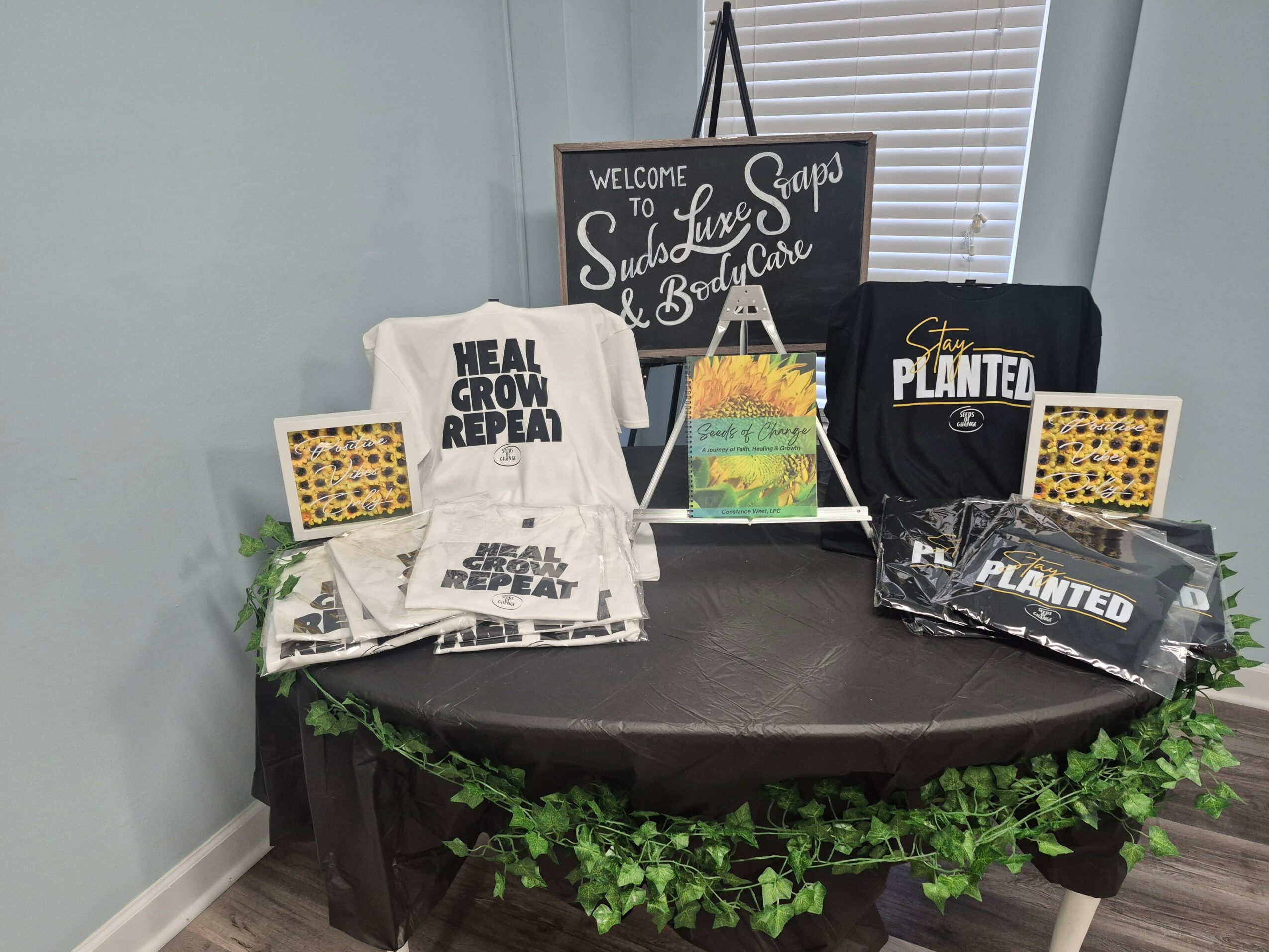 Table with various items including tote bags, framed pictures, and a chalkboard sign, decorated with green foliage around the edge.