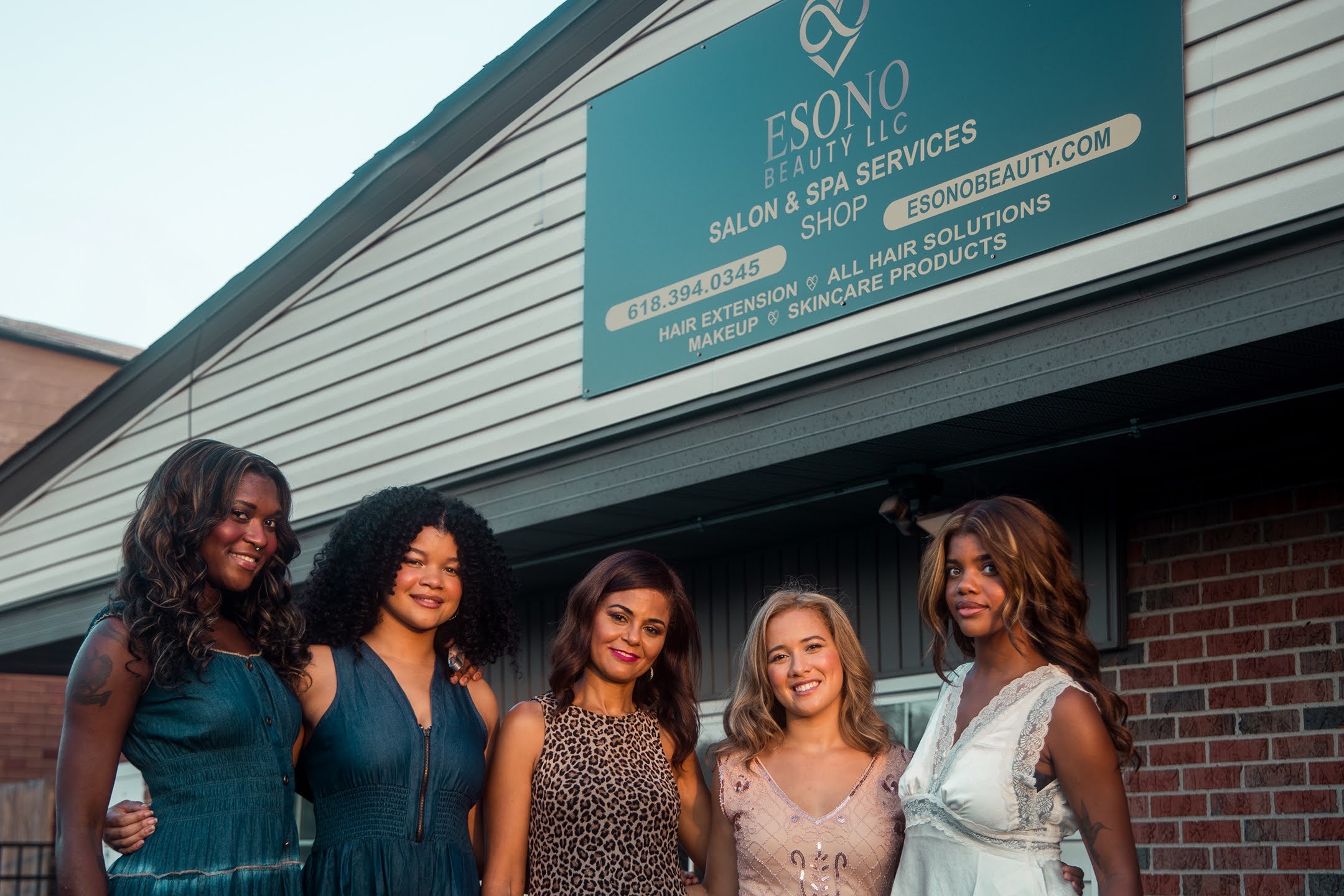 Five women standing outside a building with a sign for a salon and spa services, smiling and posing together.