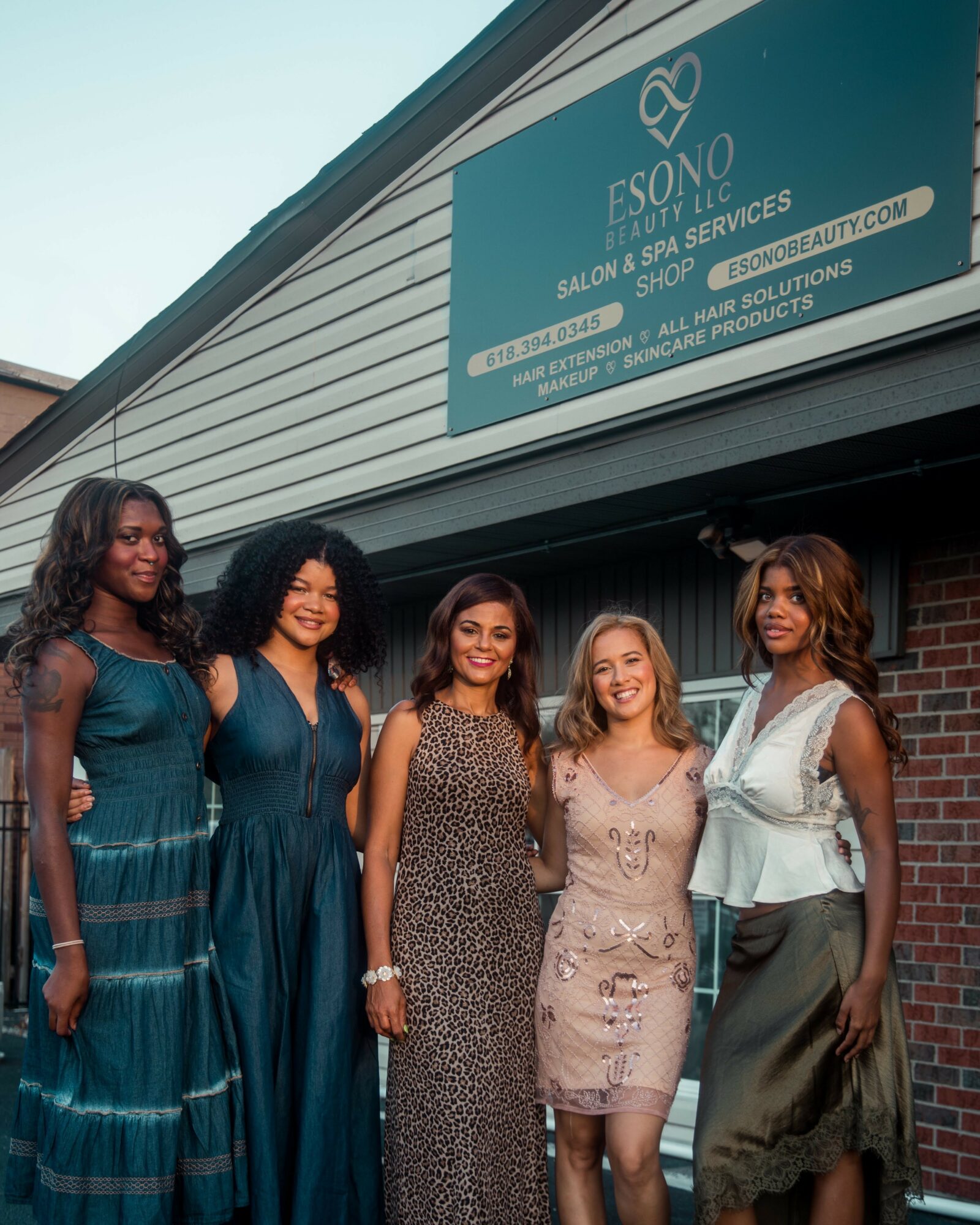 Five women stand outside a building with a sign for Esono Salon & Spa Services, smiling and posing together.