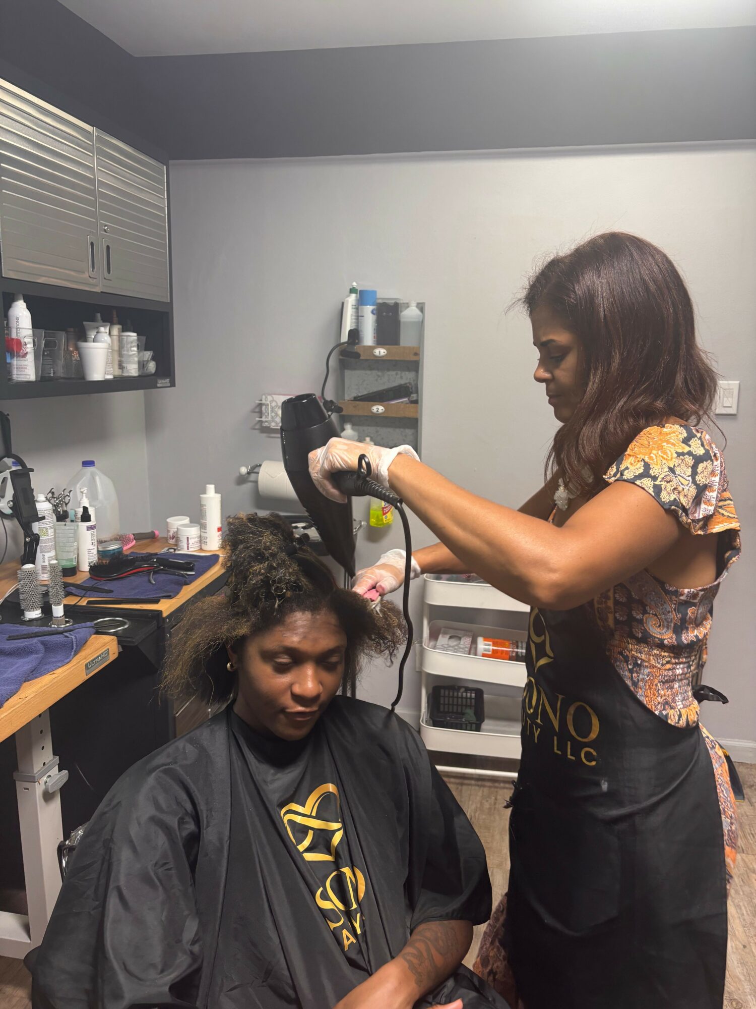 A woman with long dark hair is applying hair dye to a seated woman with curly hair in a salon.