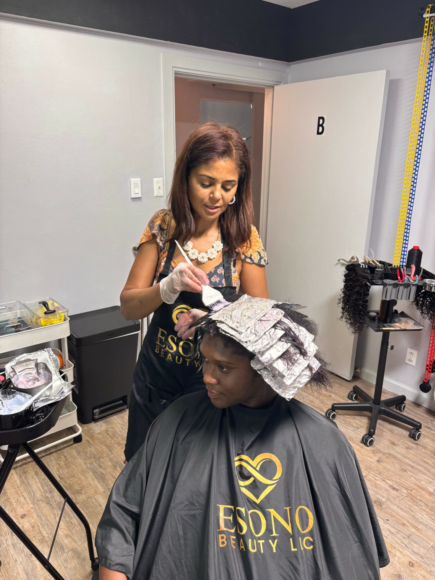 Woman getting a haircut in a salon, with hairdresser wearing gloves and cape, salon equipment visible in background.