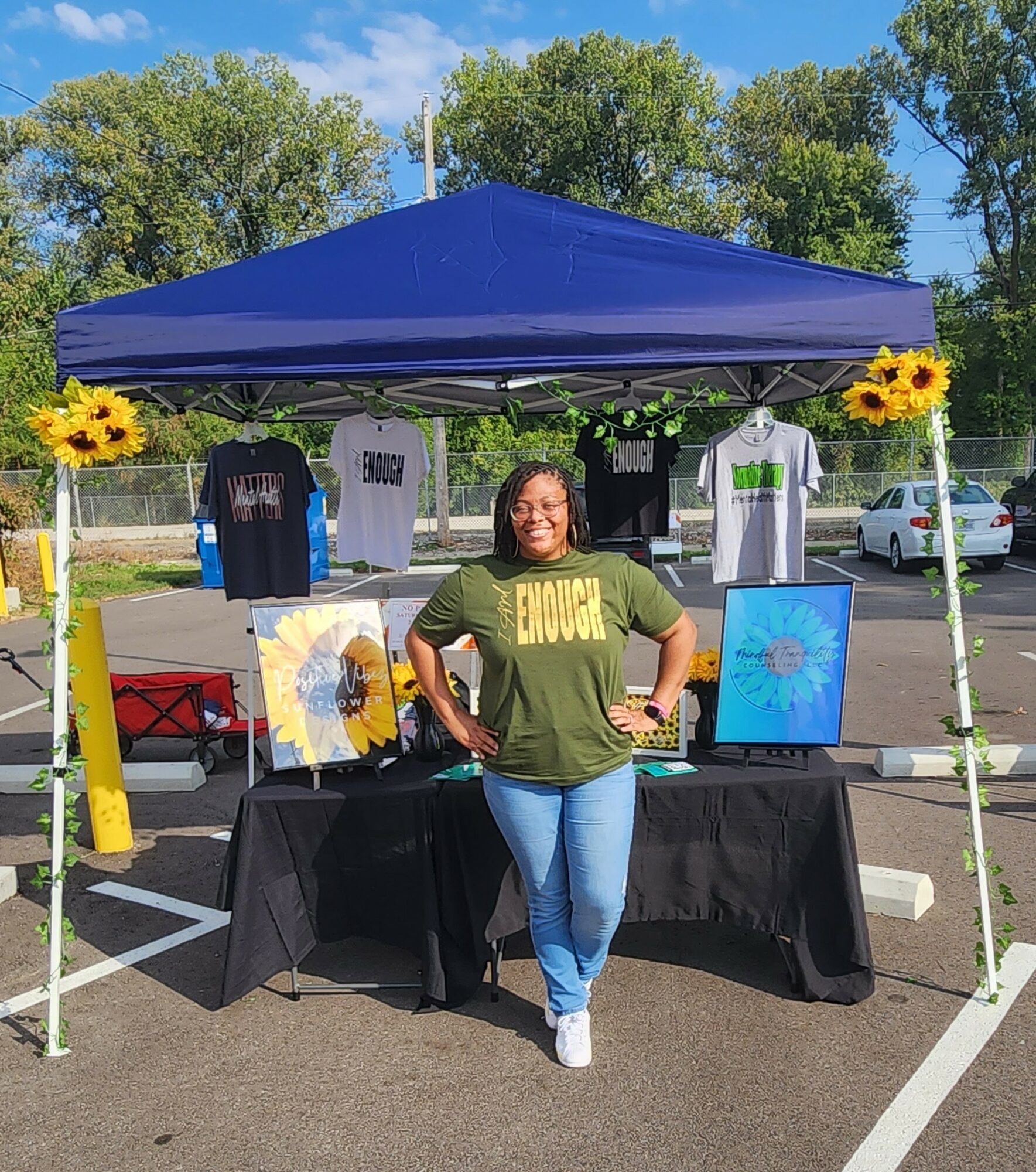 Woman standing in front of a booth with artwork and T-shirts, under a blue canopy, in a parking lot with trees.
