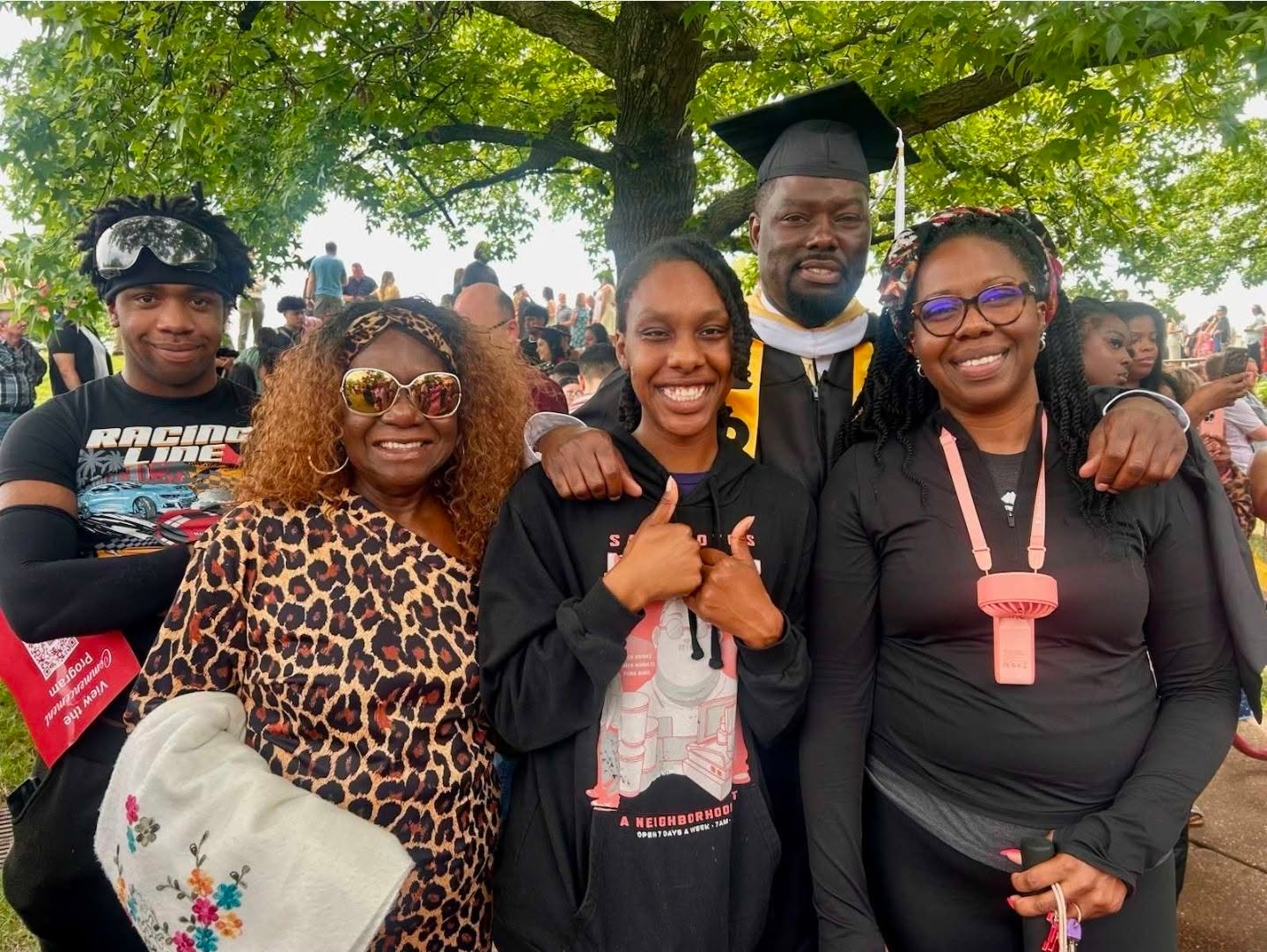 Group of four people outdoors, one in graduation gown and cap, others smiling, under a large tree.