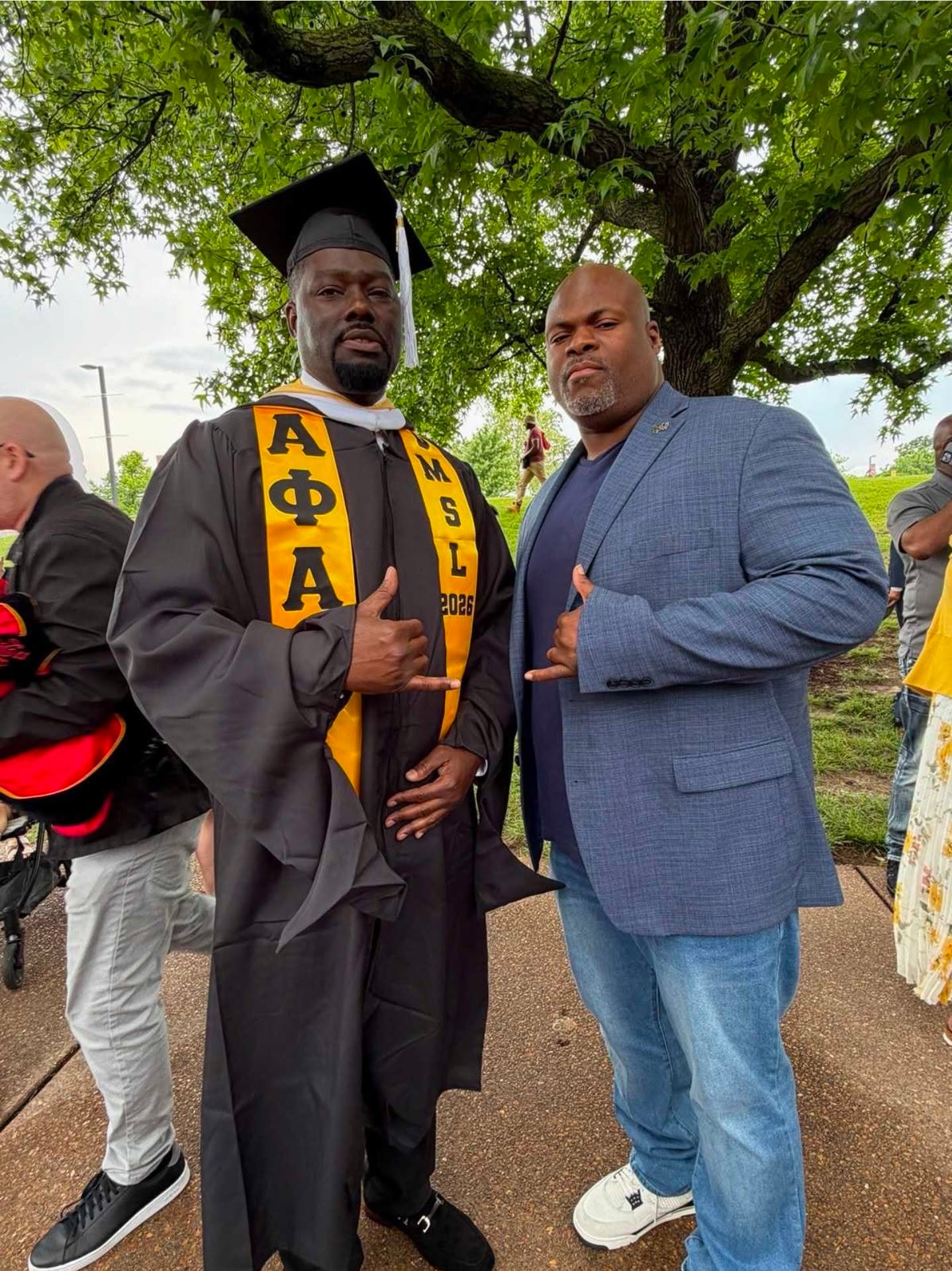 Two men standing outdoors, one in a graduation gown and cap, the other in a blue blazer, under a tree.