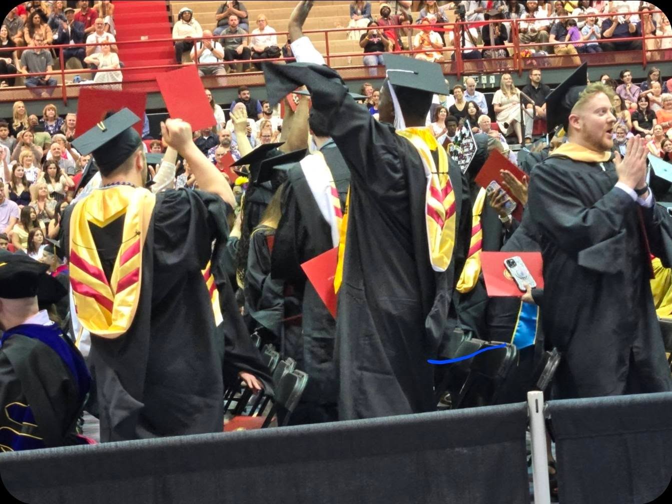 Group of graduates in caps and gowns celebrating at a graduation ceremony, with audience in the background.