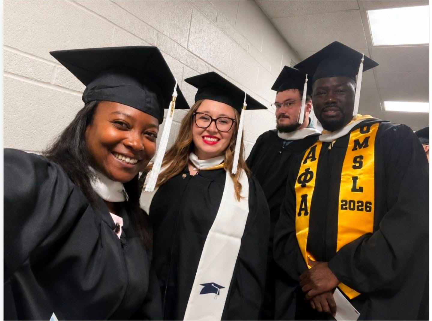Four graduates in caps and gowns smiling in a hallway, one wearing a yellow stole with Greek letters and year 2026.