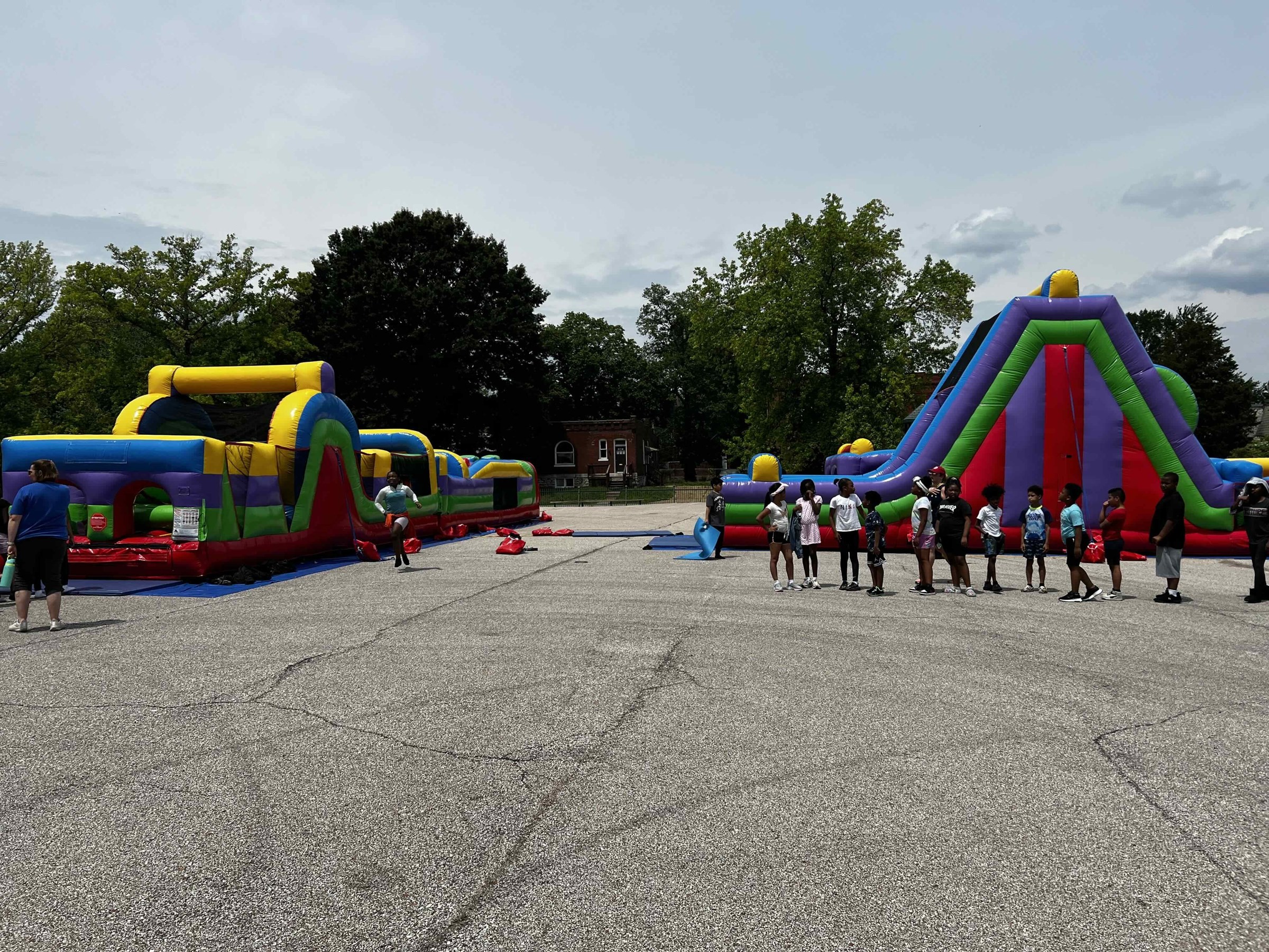 Inflatable bounce houses and slides with children playing outdoors on a cloudy day, surrounded by trees.