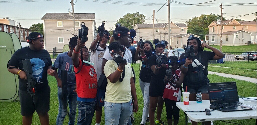 Group of people gathered outdoors, some holding guns, with a table and equipment, in a residential area.
