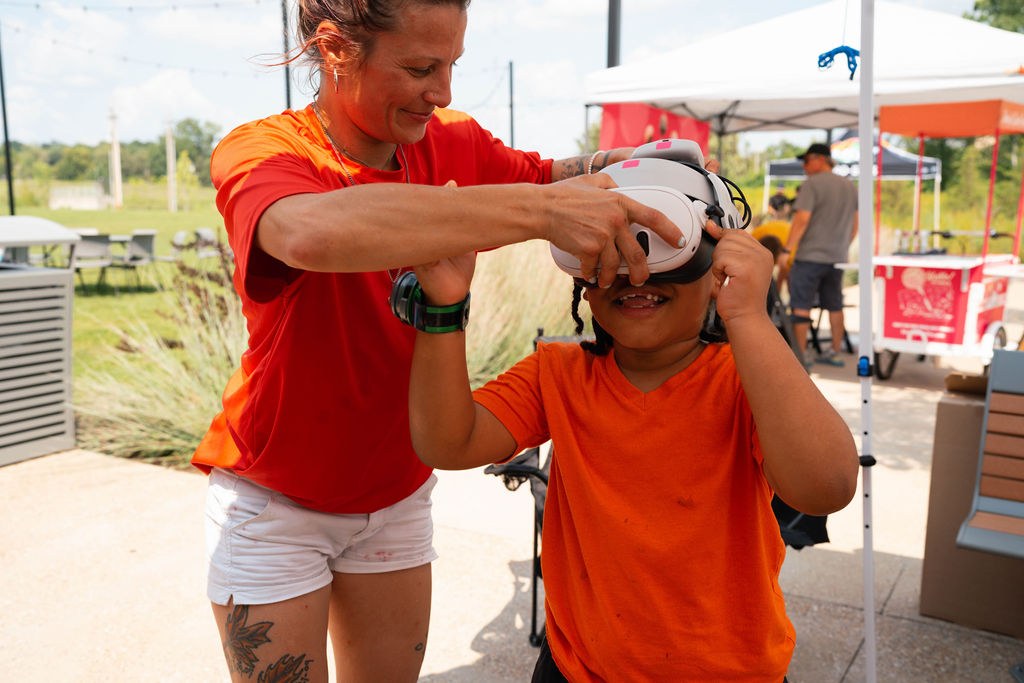 Woman helping a child put on a virtual reality headset outdoors, both wearing orange shirts.
