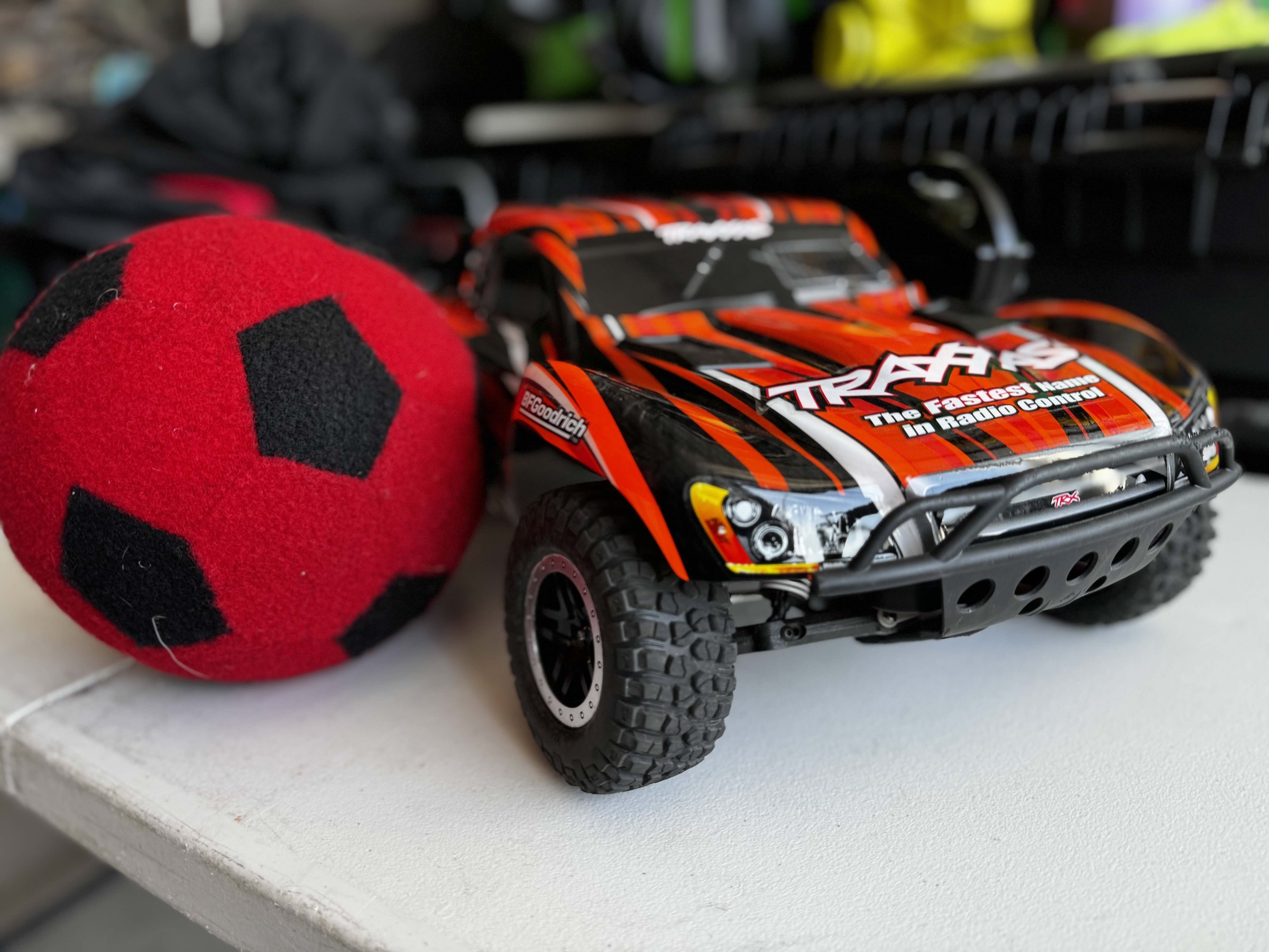 Remote-controlled car with orange and black design next to a red and black soccer ball on a white surface.