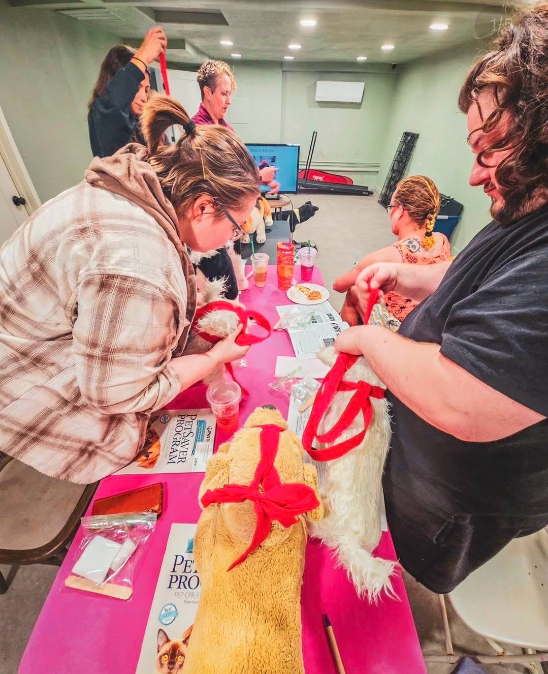 People gather around a table with craft supplies, including a stuffed animal with a red ribbon, in a room with green walls.