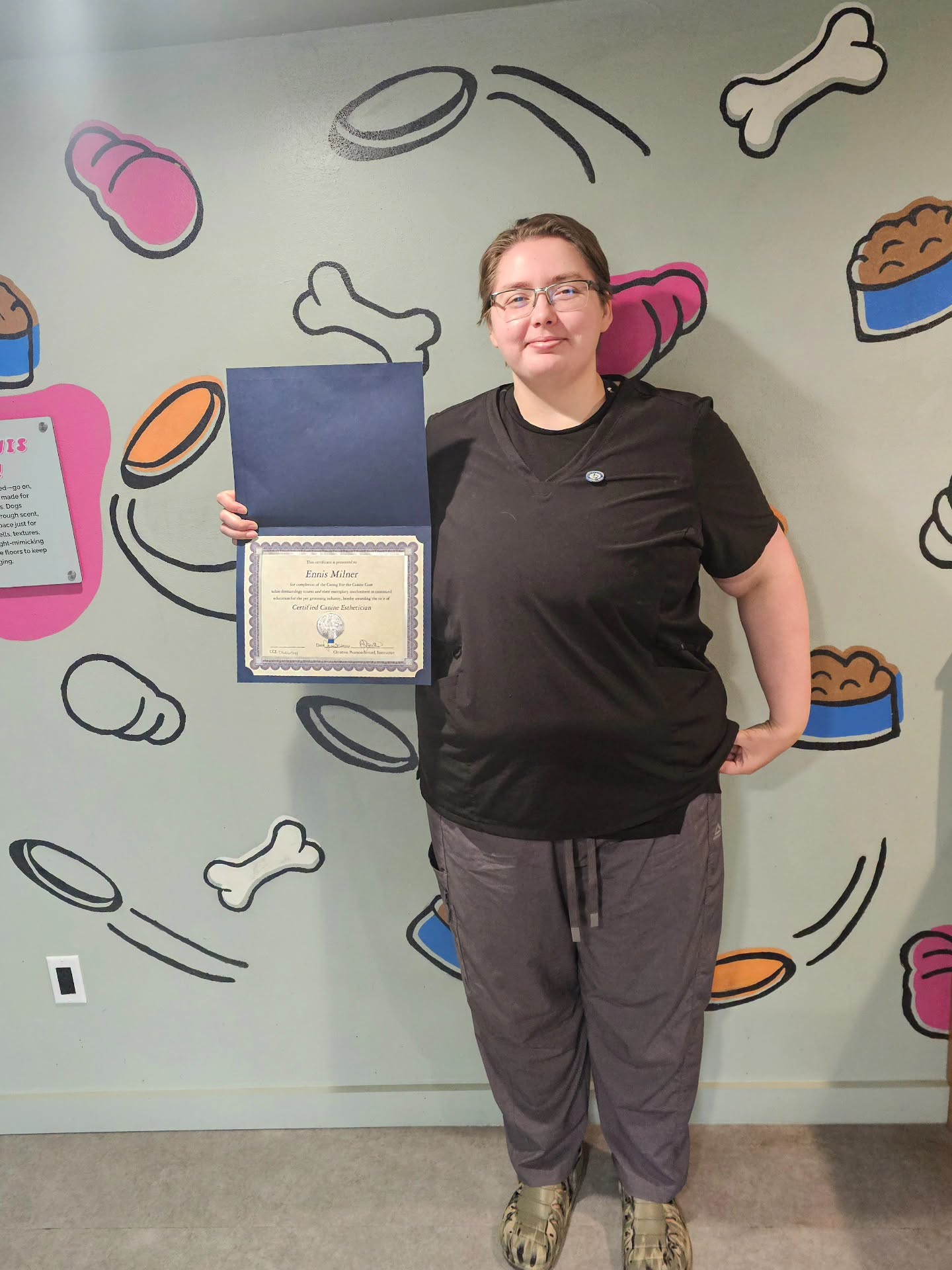 Person holding a certificate in front of a wall decorated with pet-related illustrations, smiling, wearing glasses, black shirt, gray pants.
