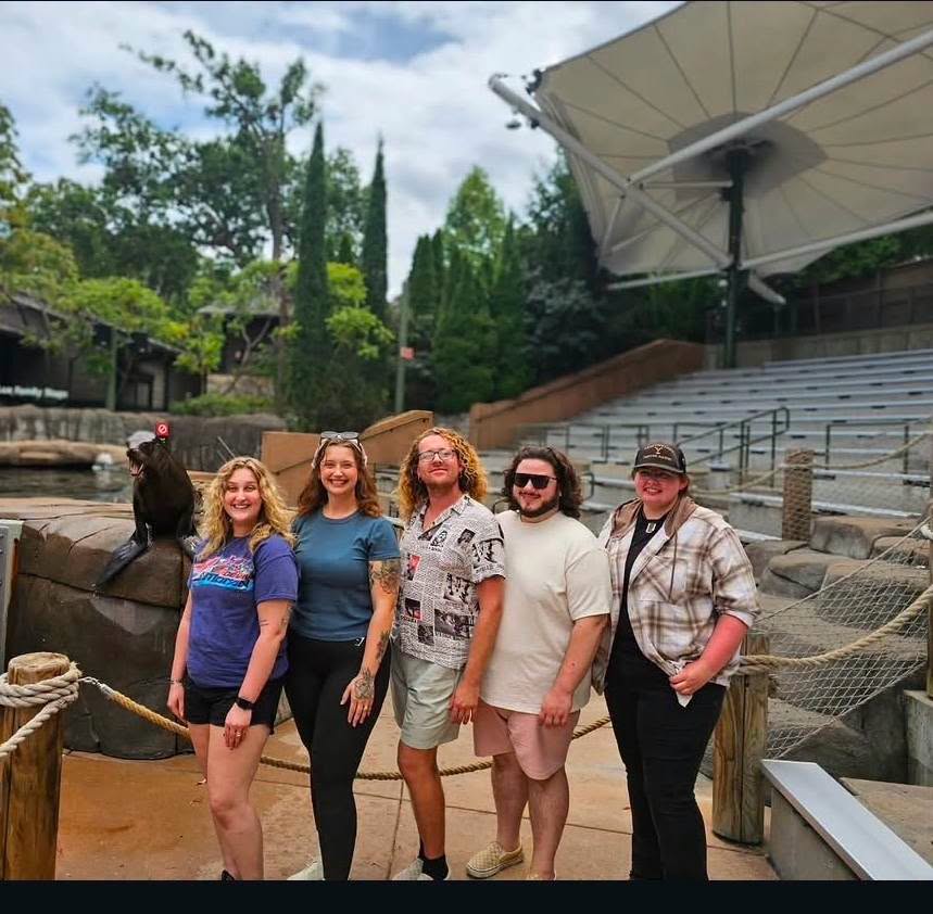Group of six people standing outdoors near a water feature and large umbrella, with trees and seating area in background.