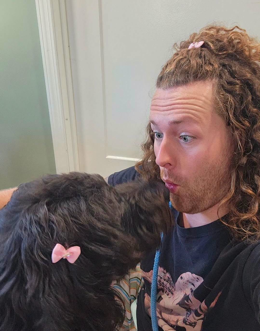 Person with curly hair and pink hair tie making a surprised face close to a black dog with pink bow, indoors.