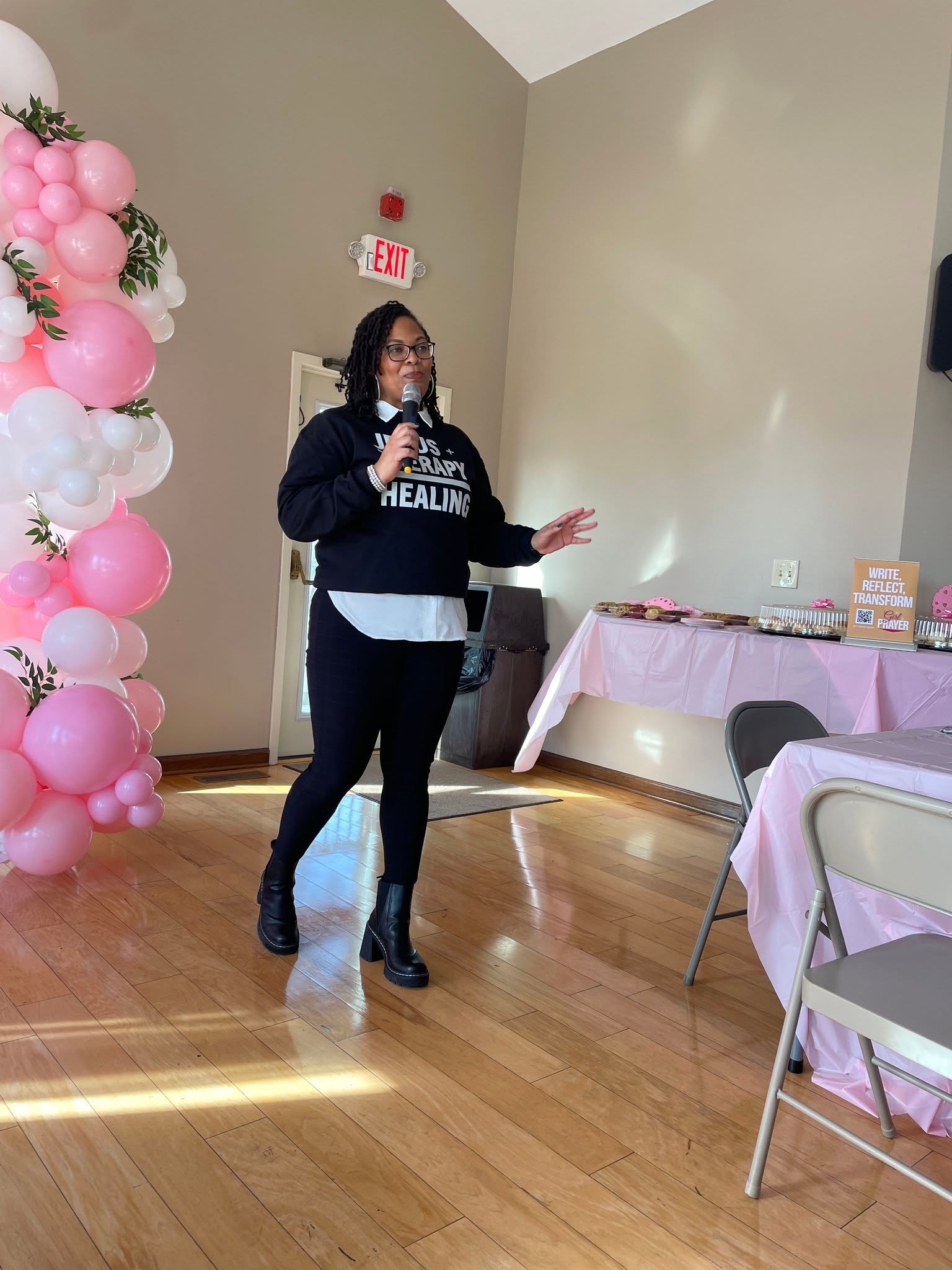 Woman speaking into microphone in a room with pink balloon decoration and tables with pink tablecloths.