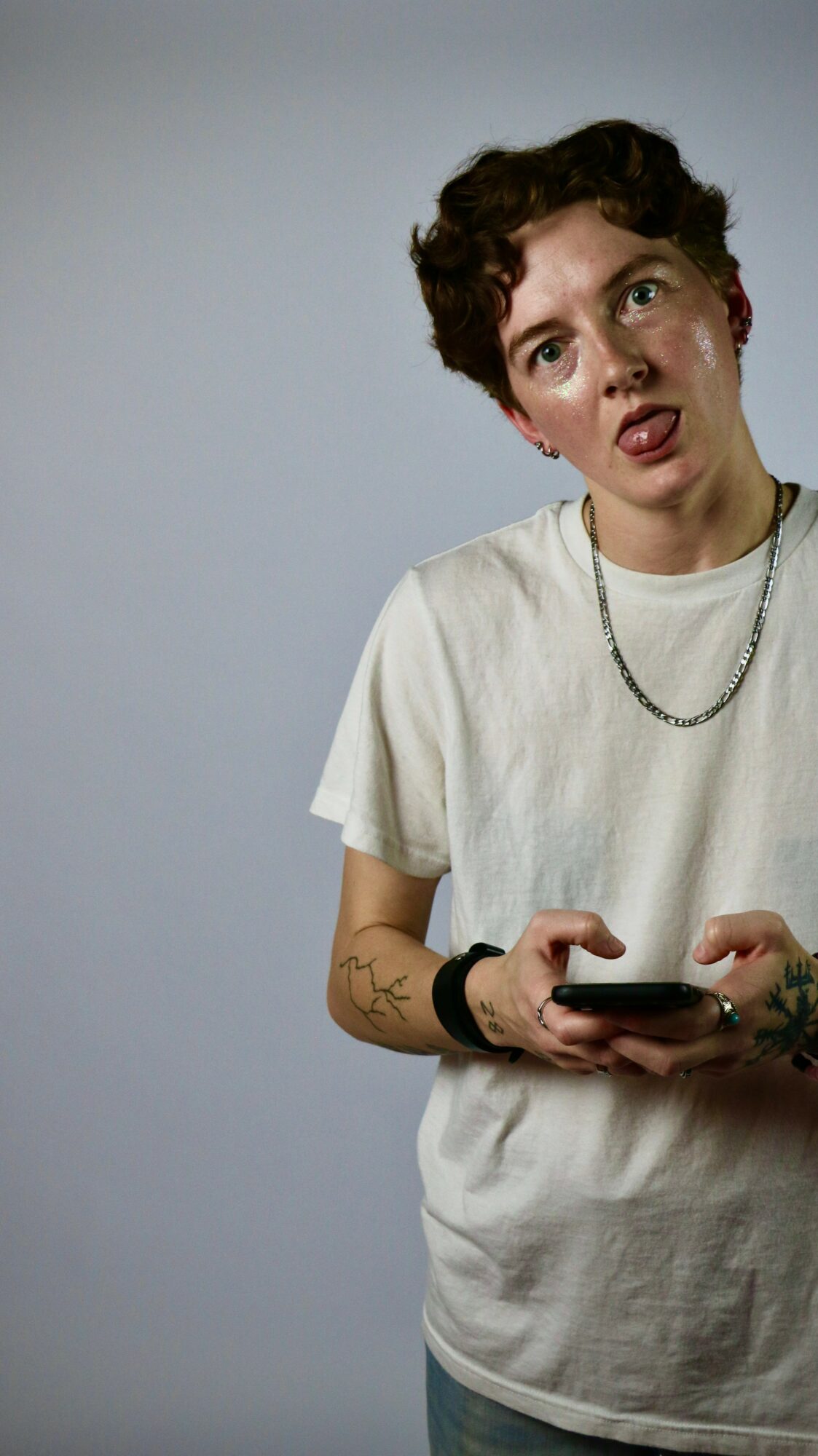 Young person with short curly hair, wearing a white t-shirt and necklace, holding a phone, looking surprised or confused.