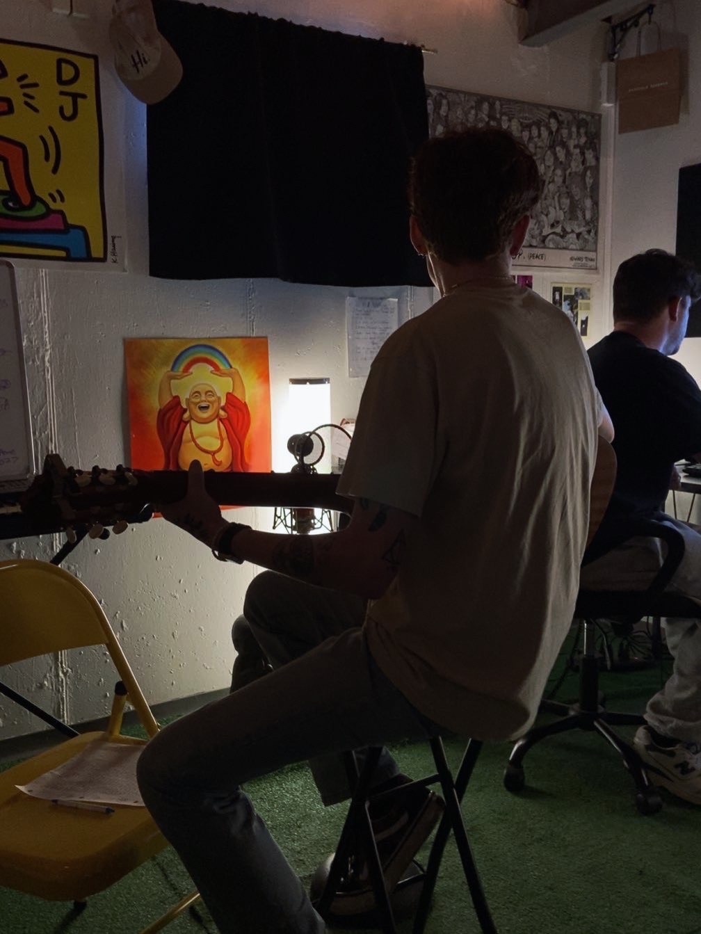 Person sitting on a stool playing guitar, facing a wall with colorful artwork, in a dimly lit room.