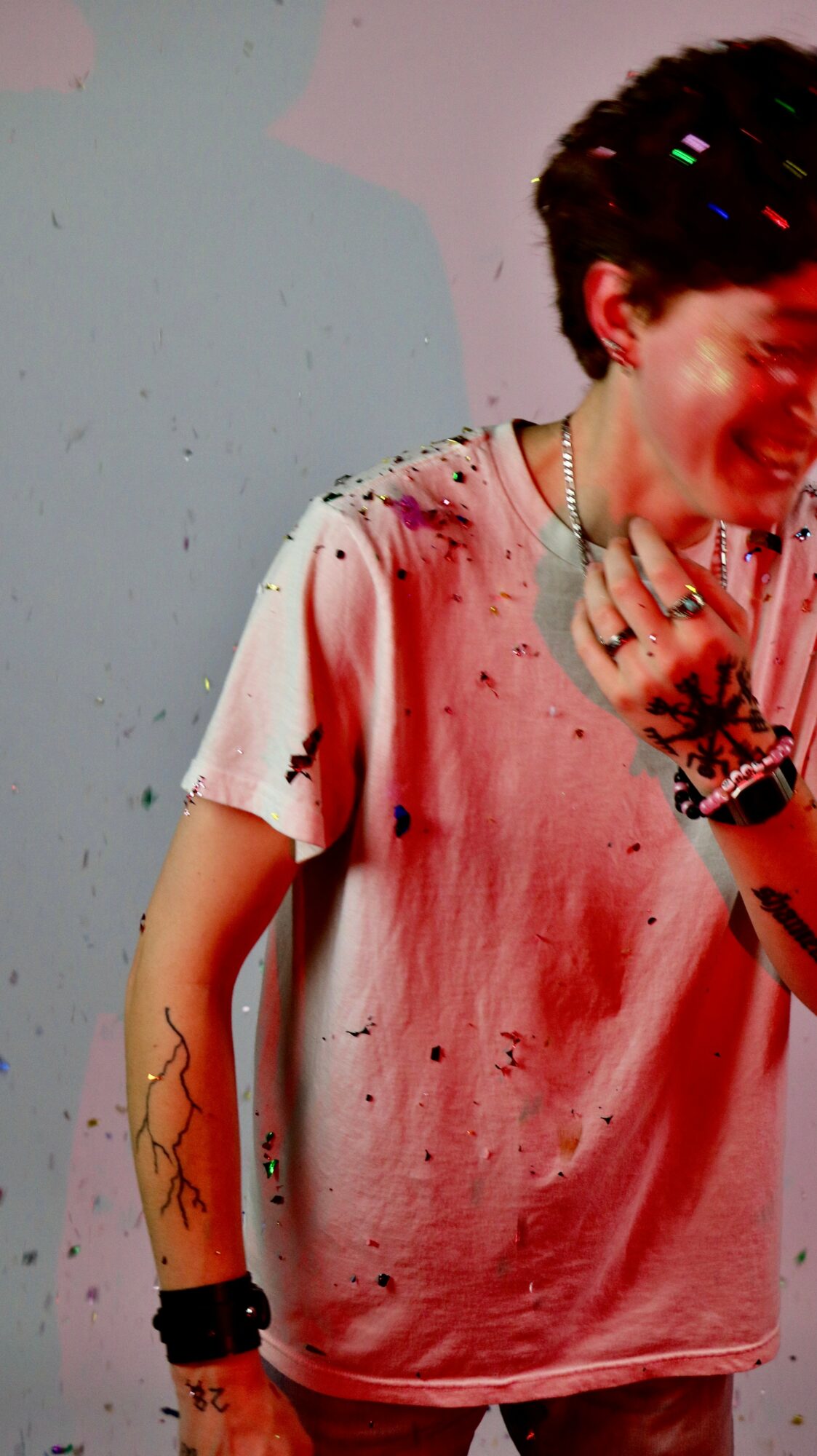 Person with short dark hair wearing a pink tie-dye shirt, smiling, with a tattoo on their arm and jewelry, against a plain background.