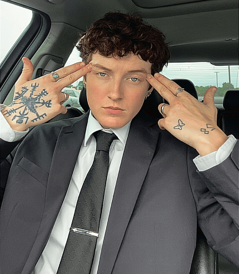 Person with short curly hair in a suit and tie, sitting in a vehicle, touching temples with both hands, tattoos visible on hands
