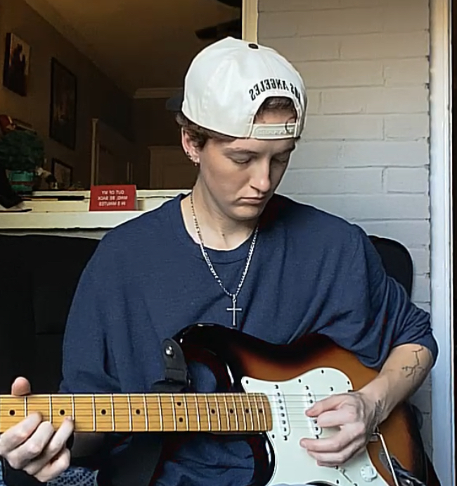 Young person wearing a white cap, playing an electric guitar indoors, with a brick wall background.
