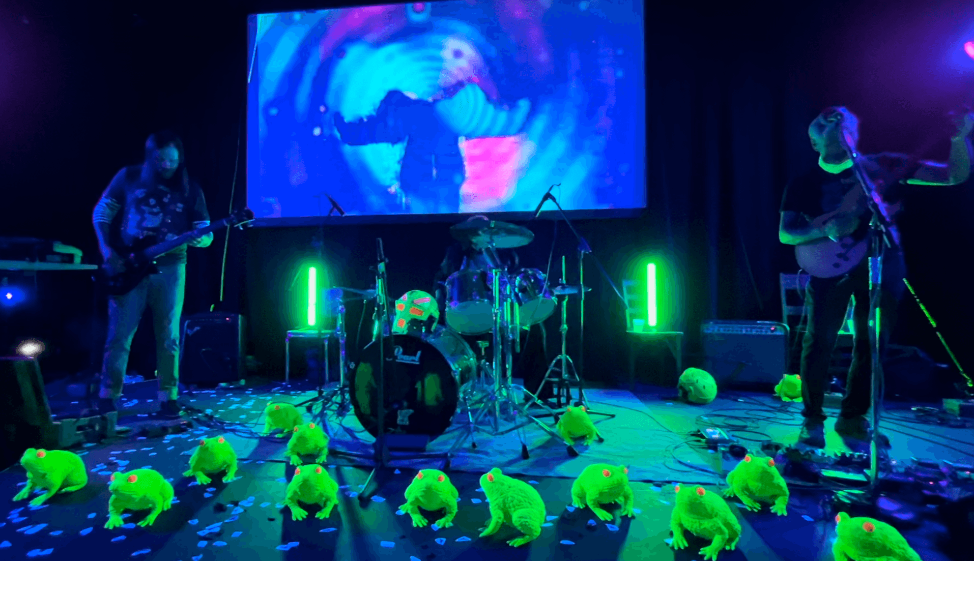 Band performing on stage with drums and guitars, colorful lighting, and a large screen in the background, with yellow plush toys on the floor.