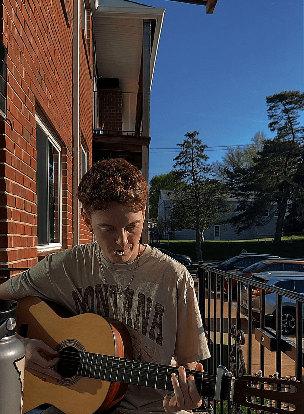 Young person playing guitar outdoors near a brick building, sunny day with trees and parked cars in background.