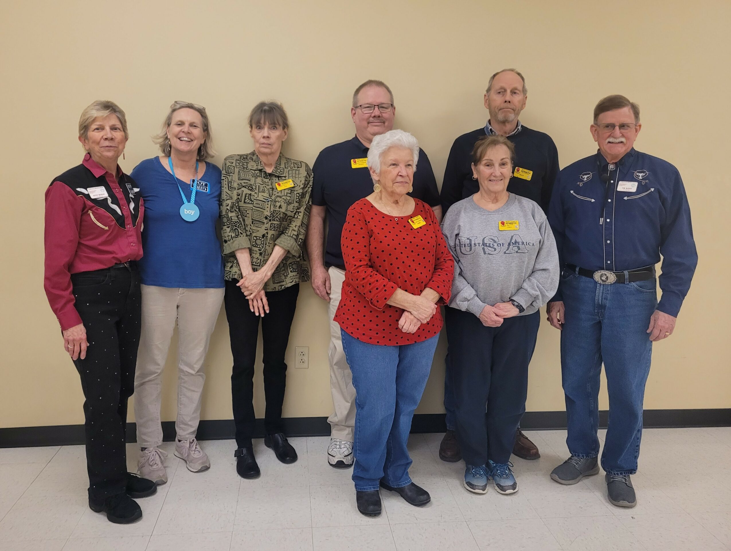 Group of eight people standing indoors against a plain wall, some wearing name tags and casual clothing.