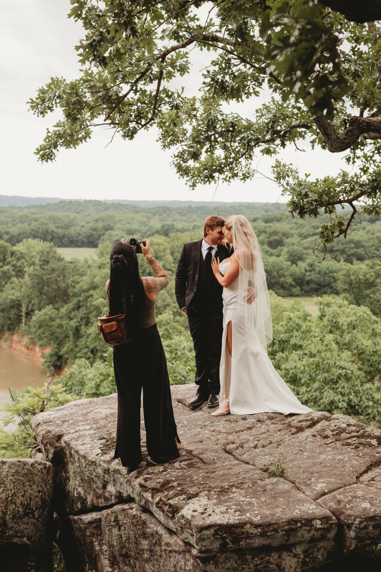 A couple in wedding attire stands on a rocky ledge outdoors, with a person taking their photo and a landscape of trees and water in the background.