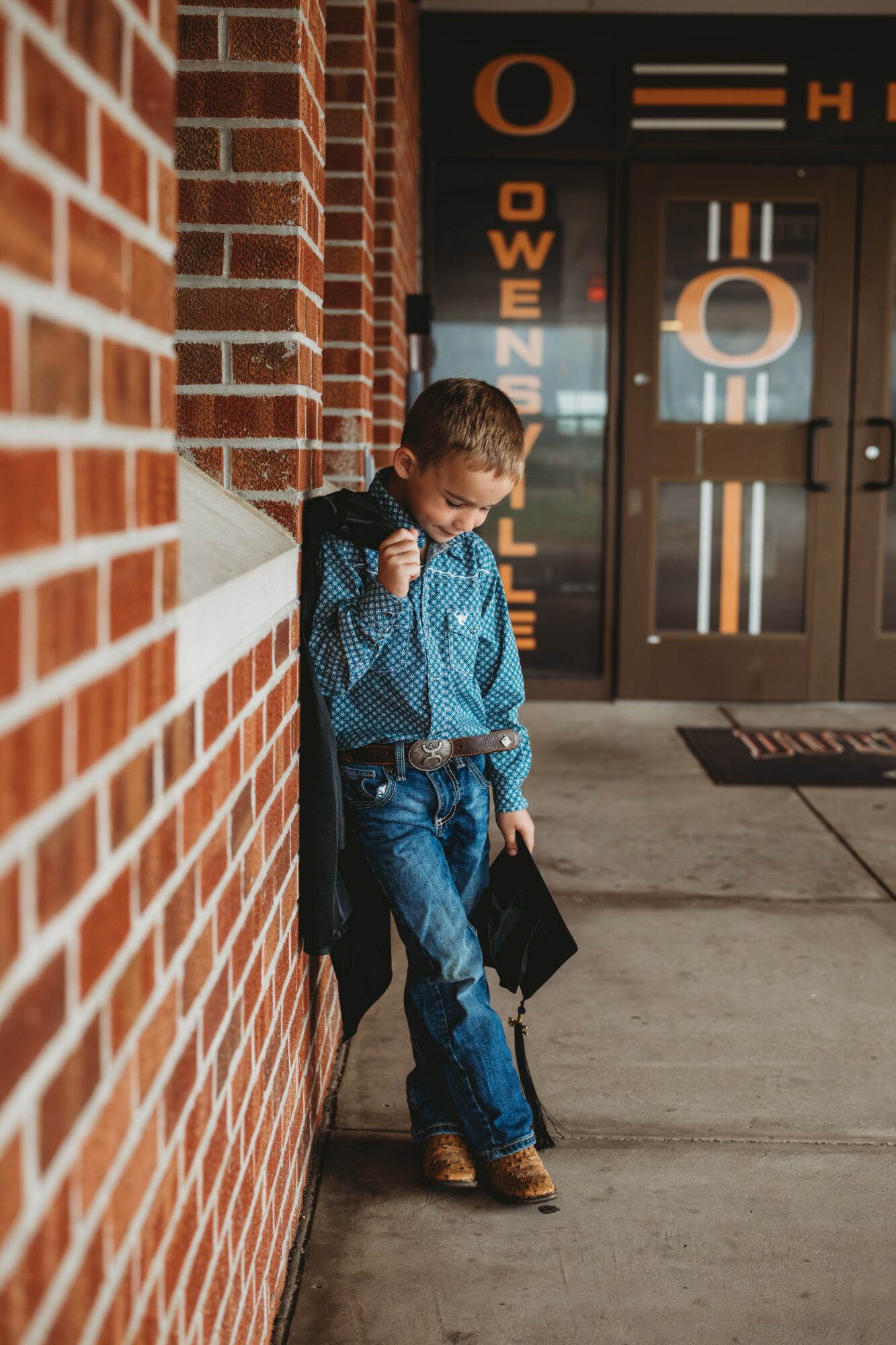 Young boy leaning against a brick wall outside a building, holding a backpack and looking down.