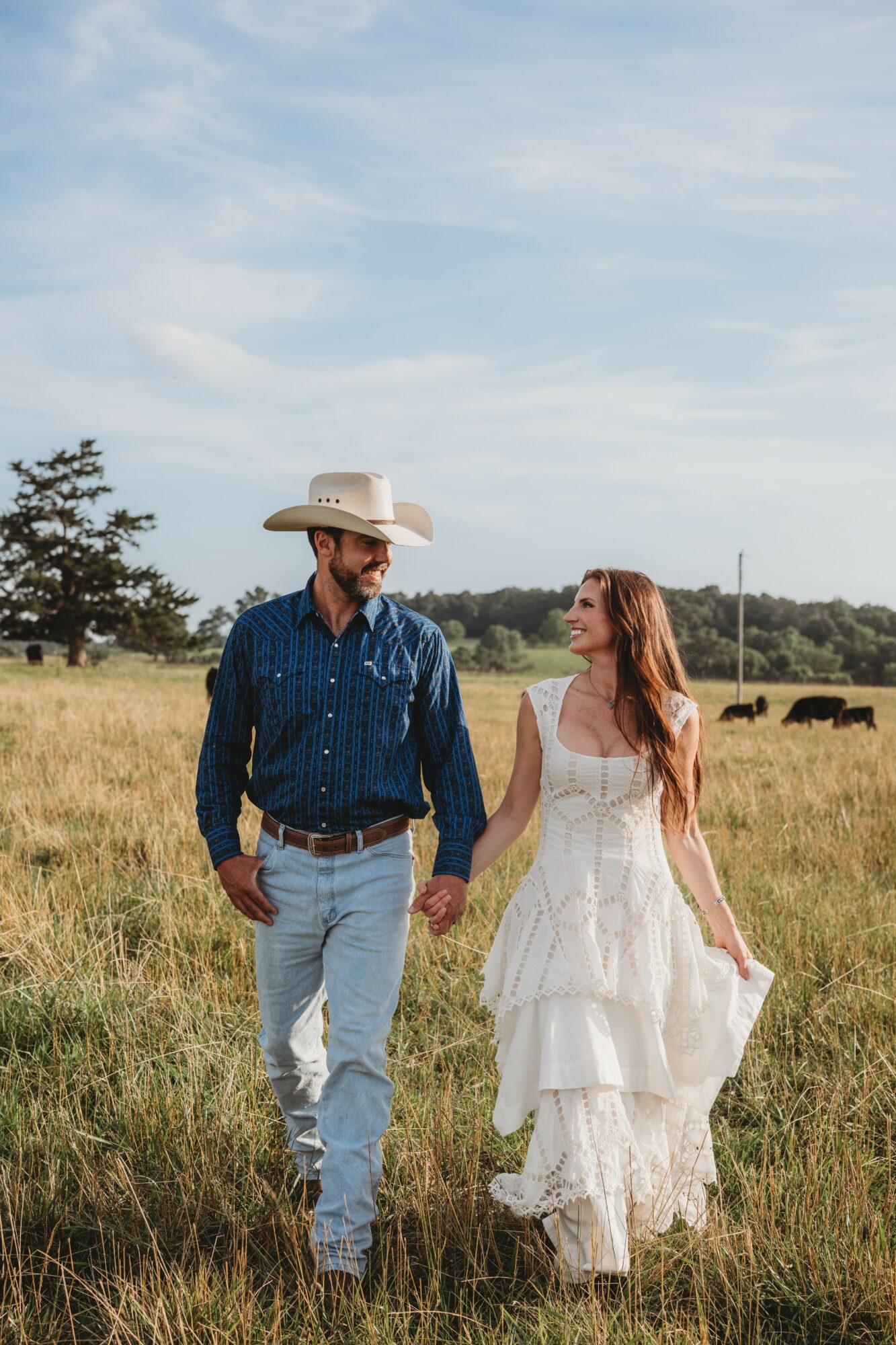 A couple holding hands walking through a grassy field under a blue sky with clouds, trees, and cattle in the background.