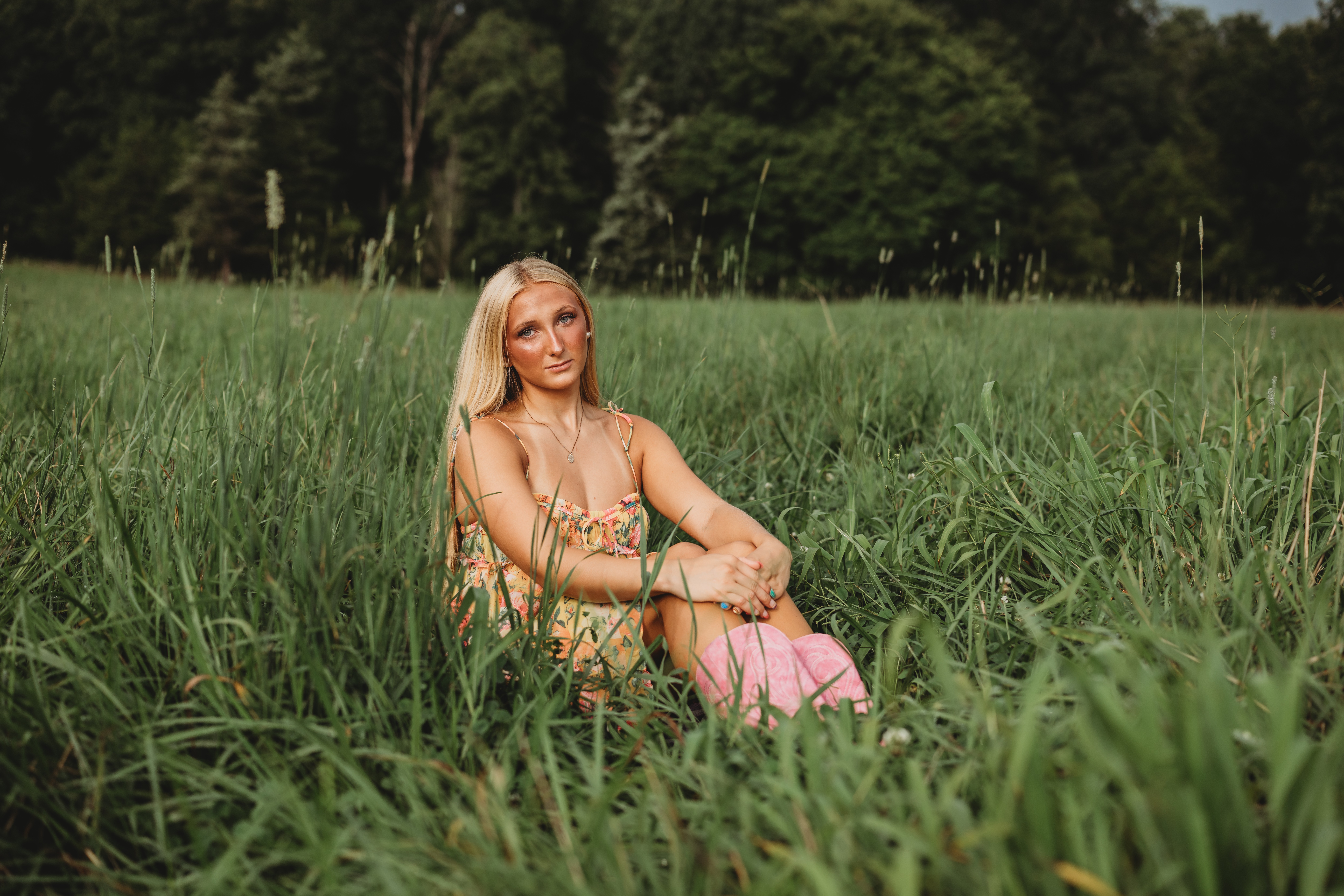 Woman sitting in tall grass in a field with trees in the background, holding a pink bag, smiling.