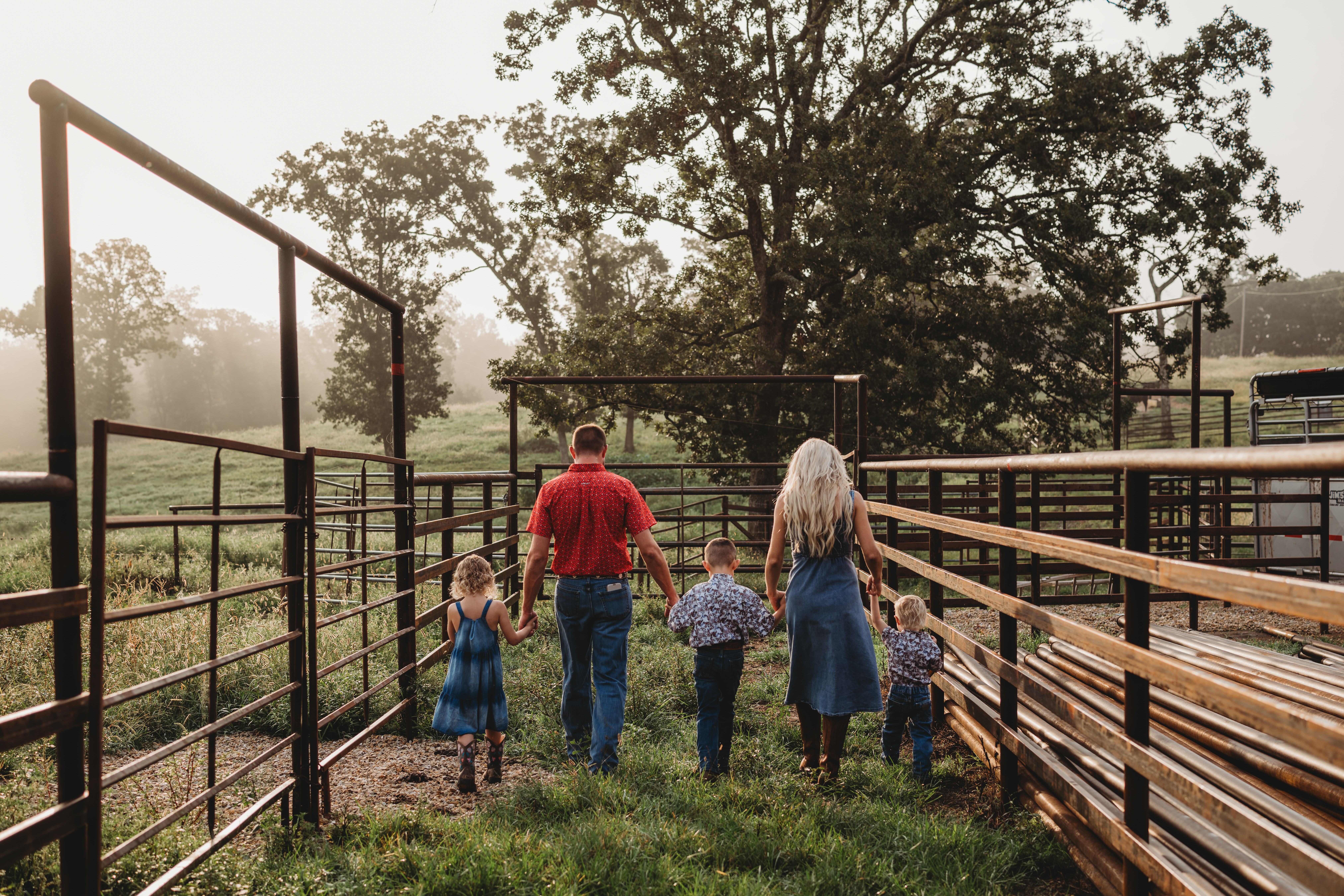 Family of five walking away on a farm path, with trees and metal fencing on both sides, overcast sky.