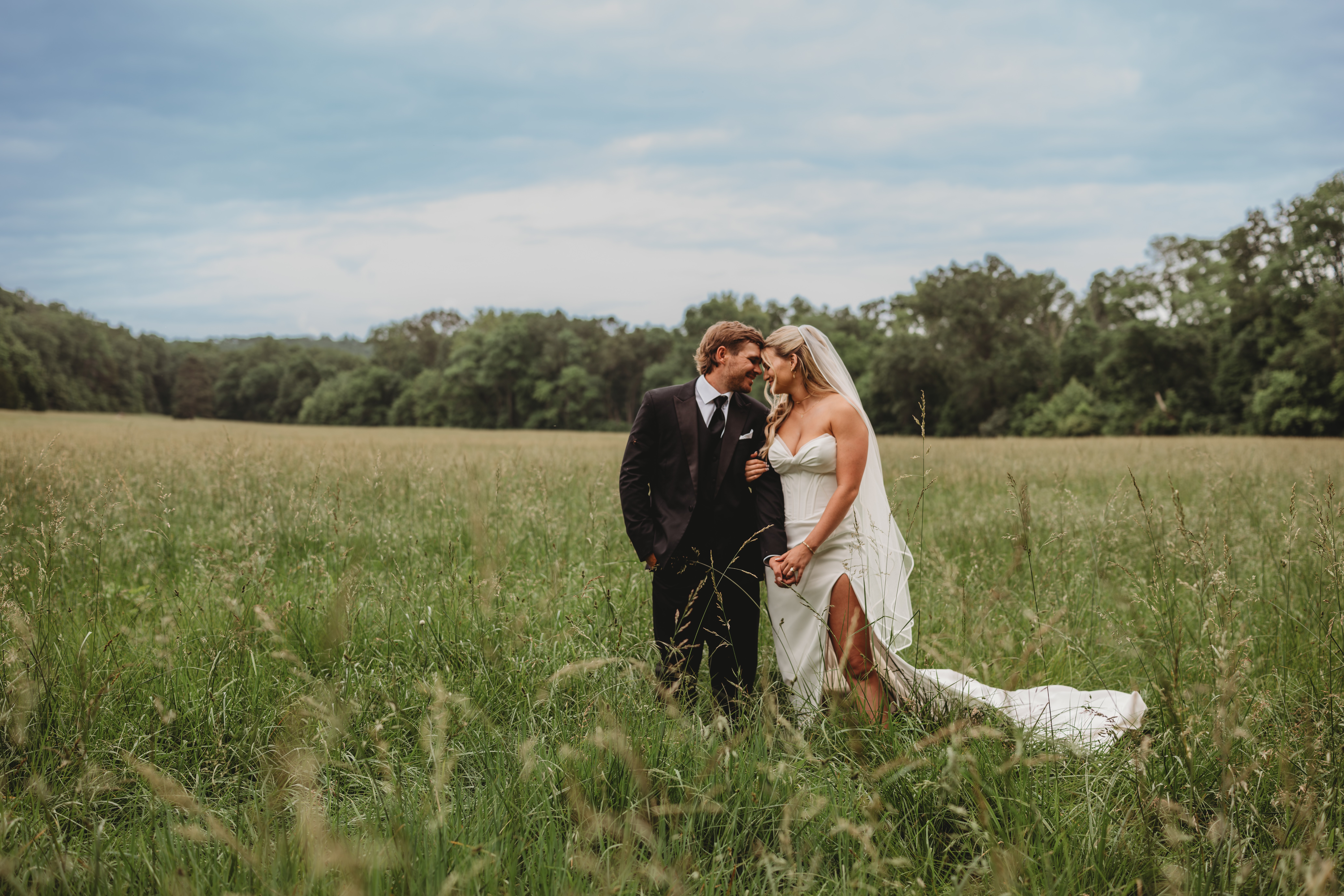 Bride and groom standing close in a grassy field, with trees and sky in the background.