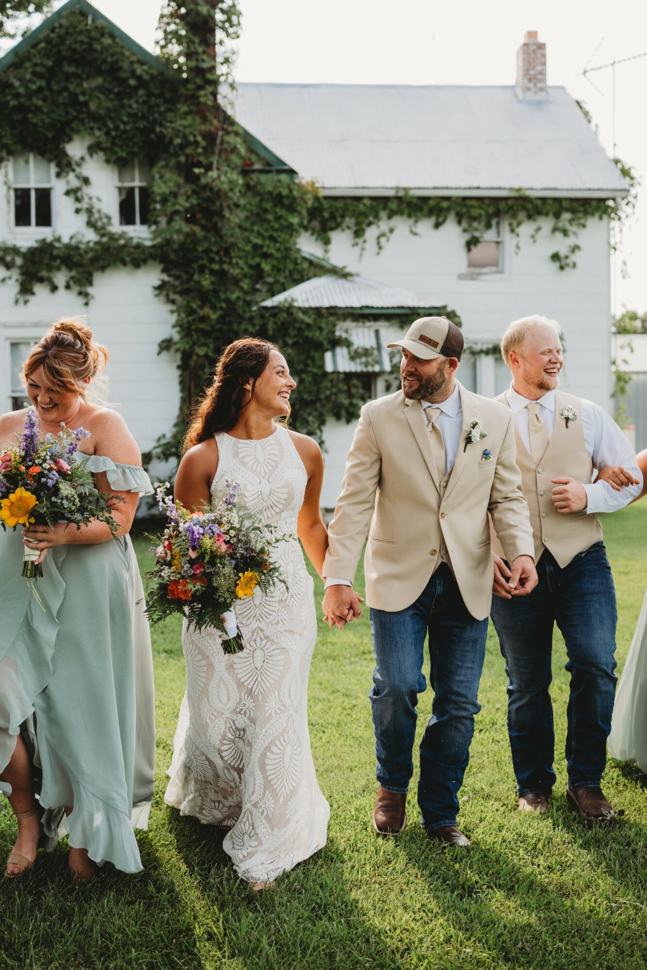 Four people walk outdoors in front of a white house with greenery, two women hold bouquets, two men smile and talk.