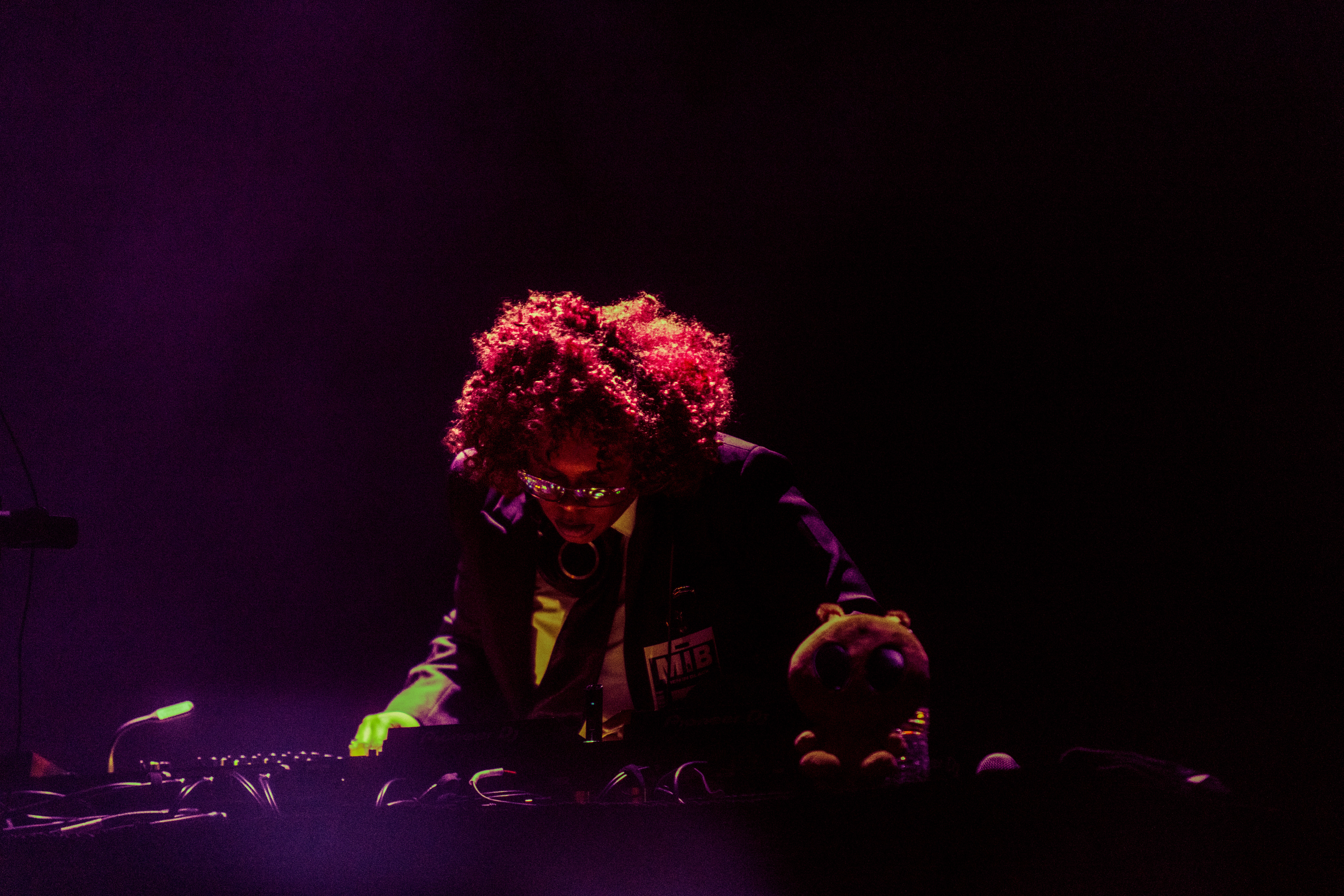 Person with curly hair wearing glasses and a dark jacket, working with electronic equipment in a dark setting.