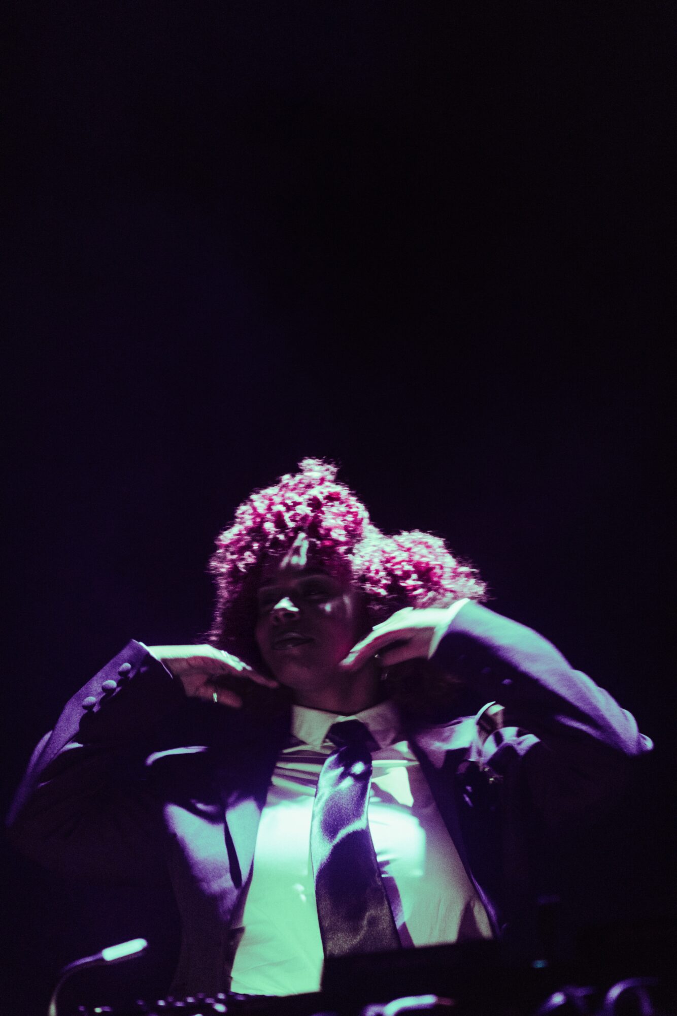 Person with curly hair adjusting glasses in a dark setting, wearing a jacket and tie.