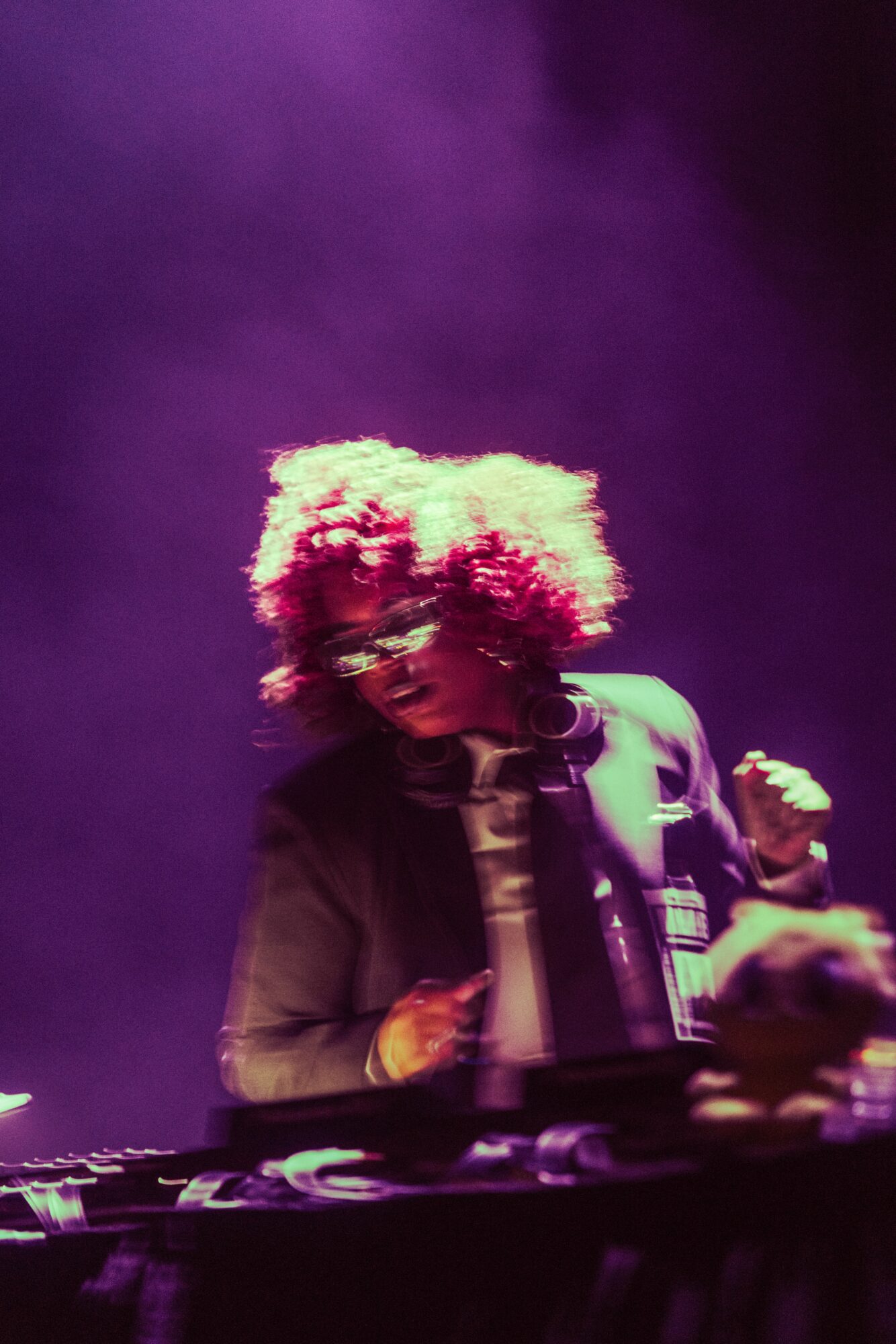 Person with curly hair wearing glasses and a jacket, performing on stage with purple lighting